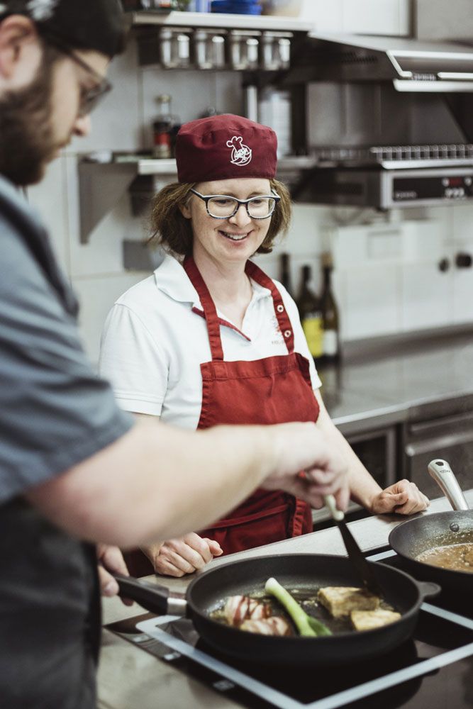 Zwei Köche in einer professionellen Küche, eine Frau in roter Schürze und Mütze lächelt, während ein Mann in einer Pfanne kocht.