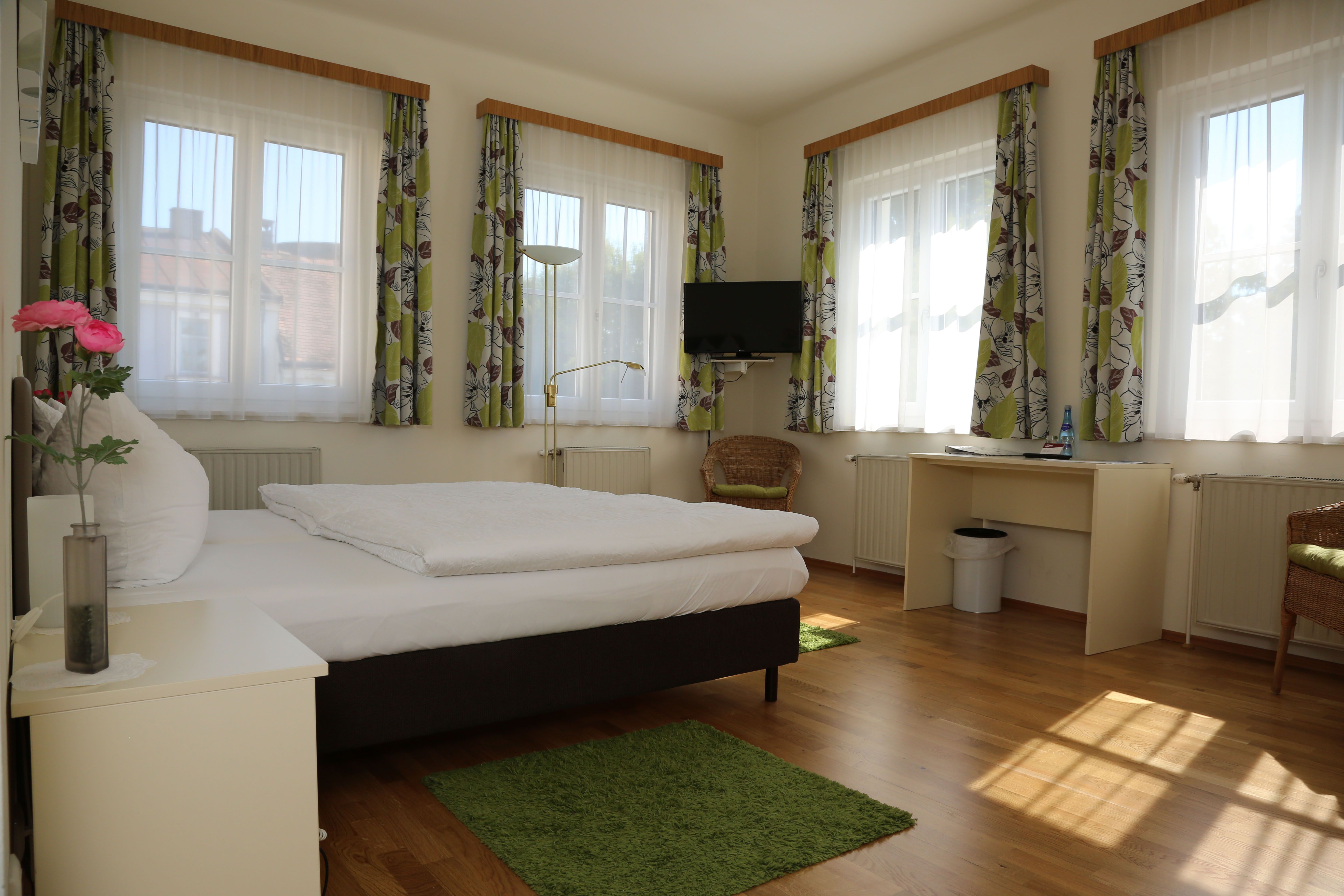 Bright room with bed, desk, TV and several windows with curtains.