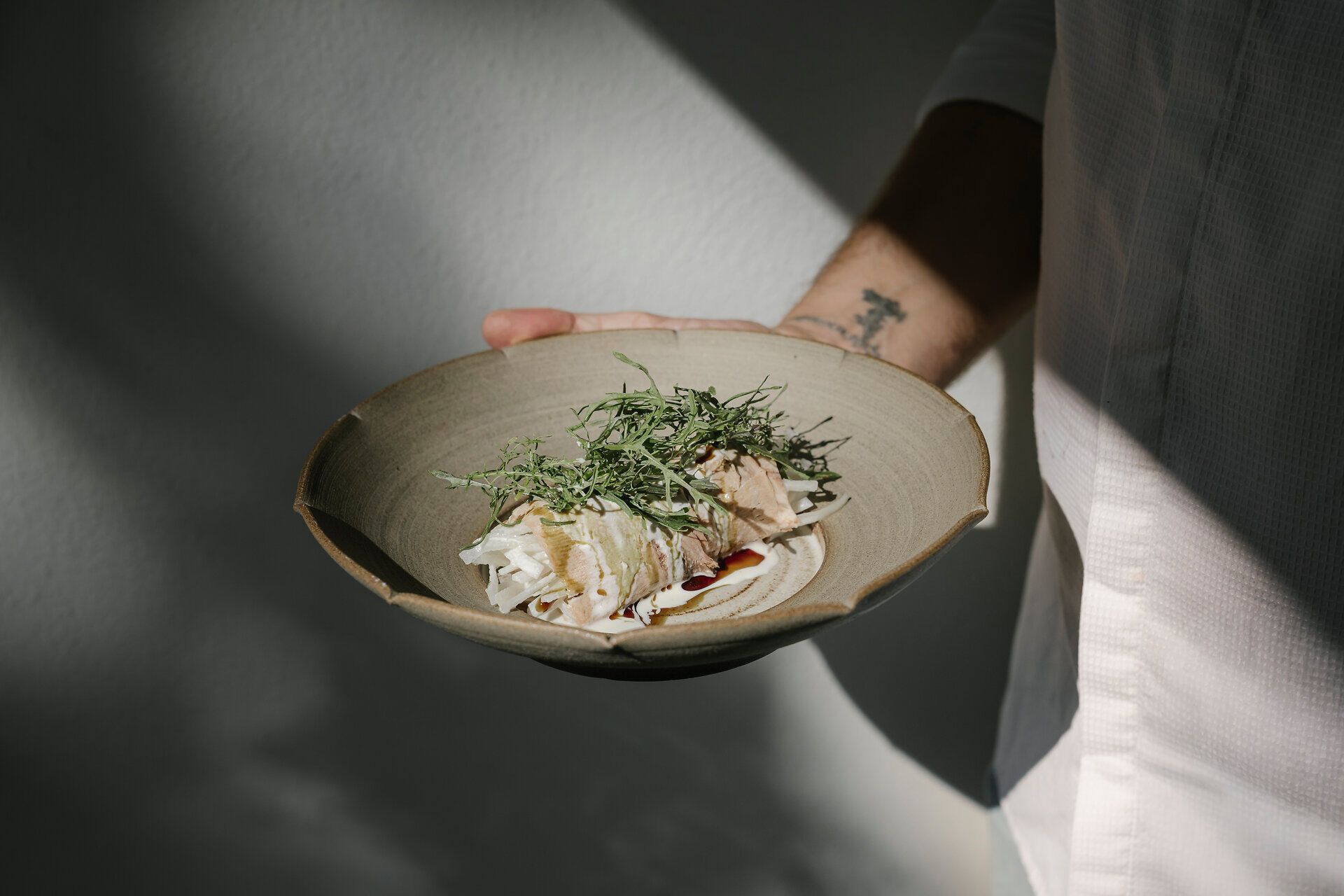 Eine Person hält einen Keramikteller mit einem angerichteten Gericht aus Fleisch, fein geschnittenem Gemüse, grünen Kräutern und Sauce in den Händen.