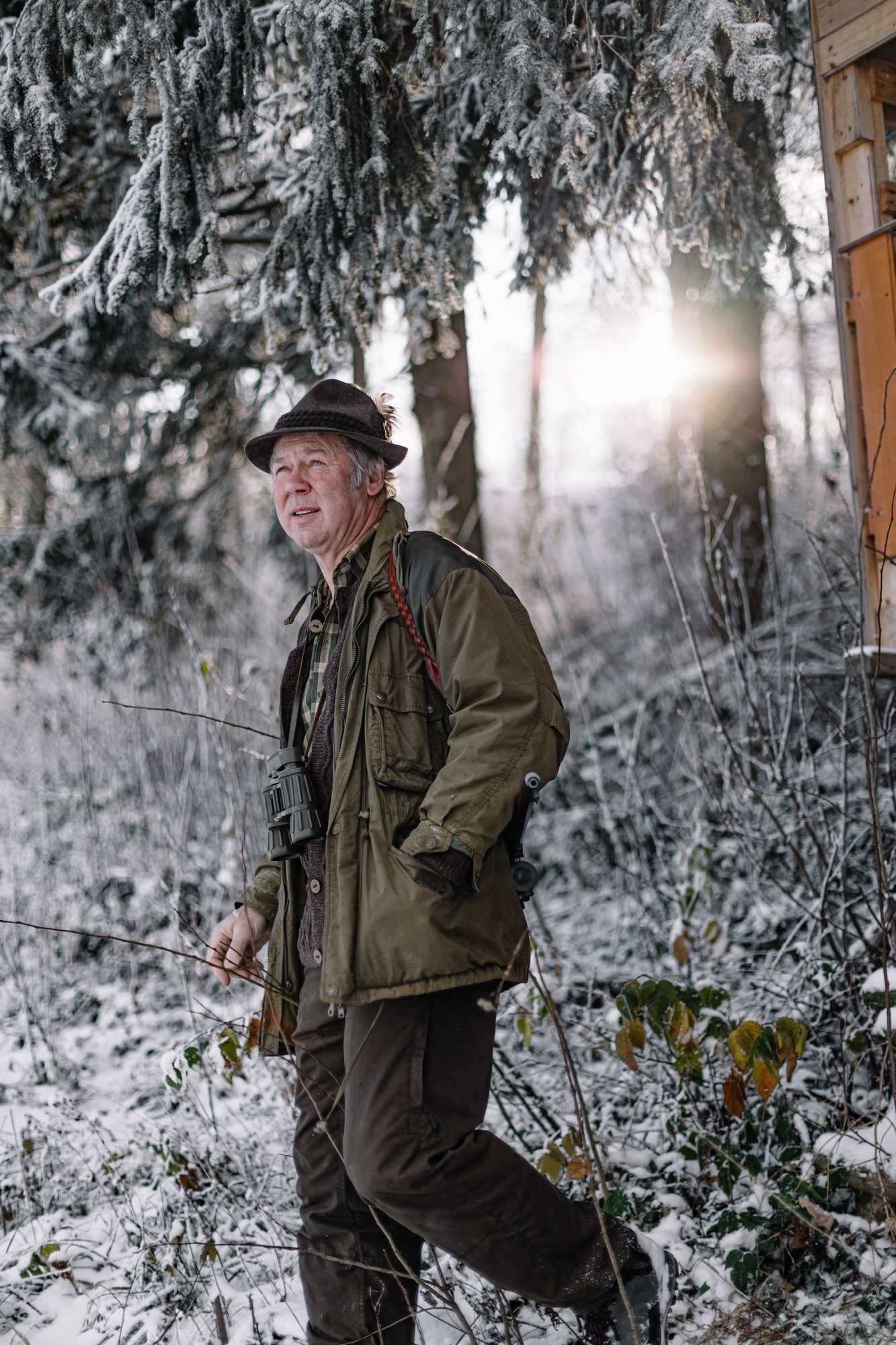 Ein älterer Mann in Jagdkleidung steht im verschneiten Wald mit Fernglas um den Hals.