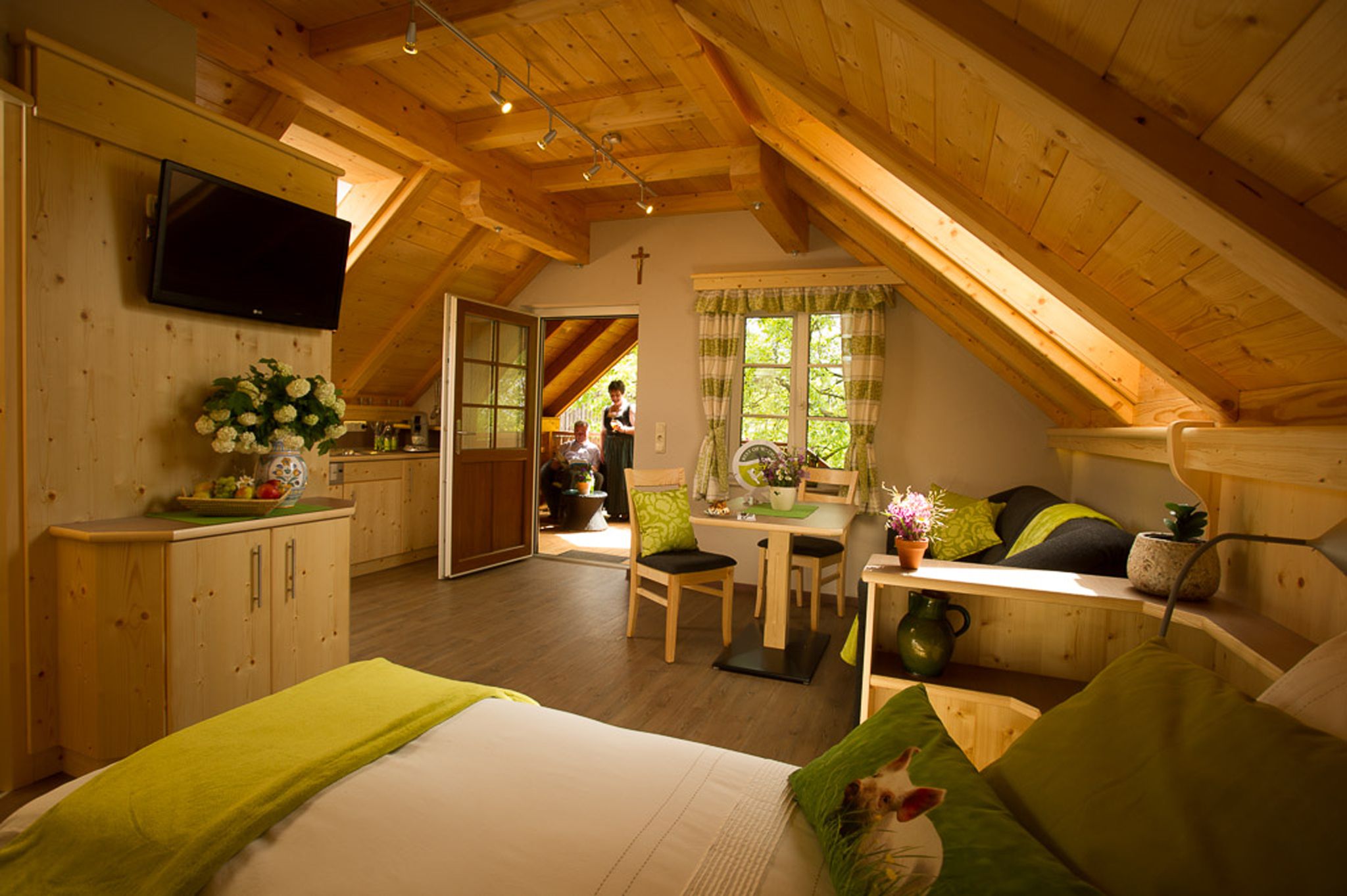 Gemütliche Ferienwohnung mit Holzausstattung, Dachschrägen und grünen Akzenten. Im Hintergrund sind zwei Personen auf einem Balkon zu sehen.