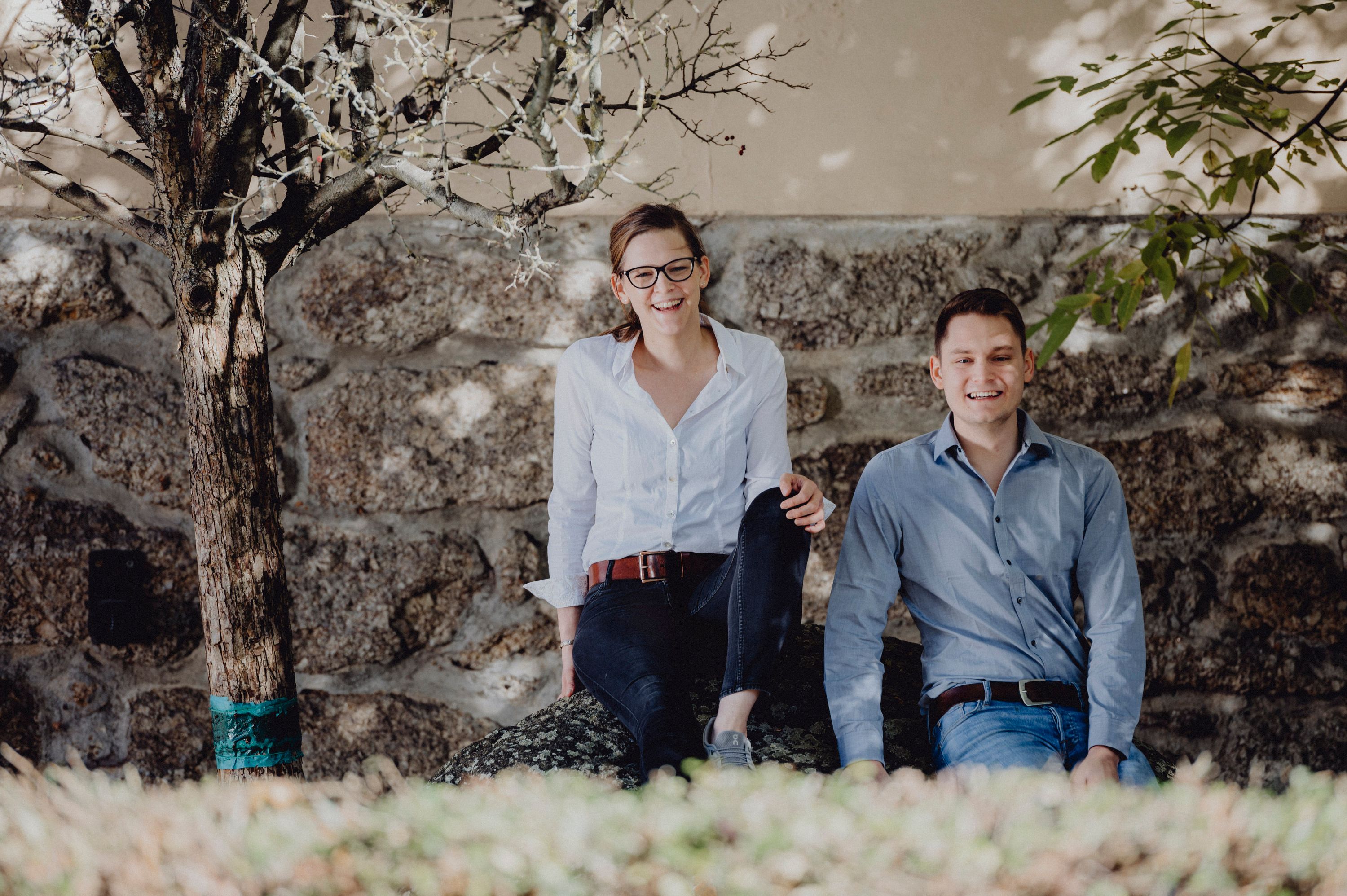 Two people sit smiling on a stone in front of a stone wall, surrounded by trees.
