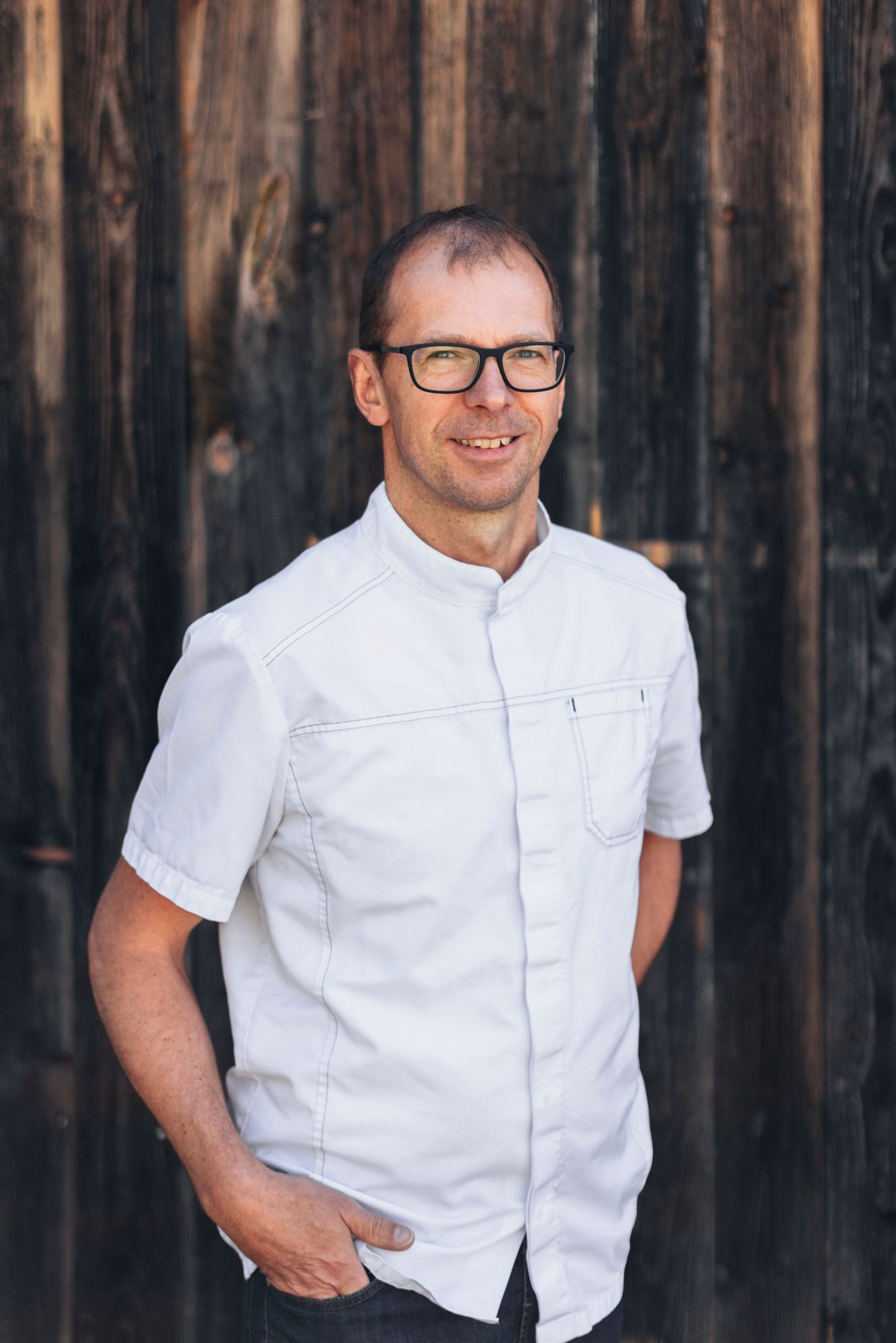Mann mit Brille und weißem Hemd vor Holzwand.