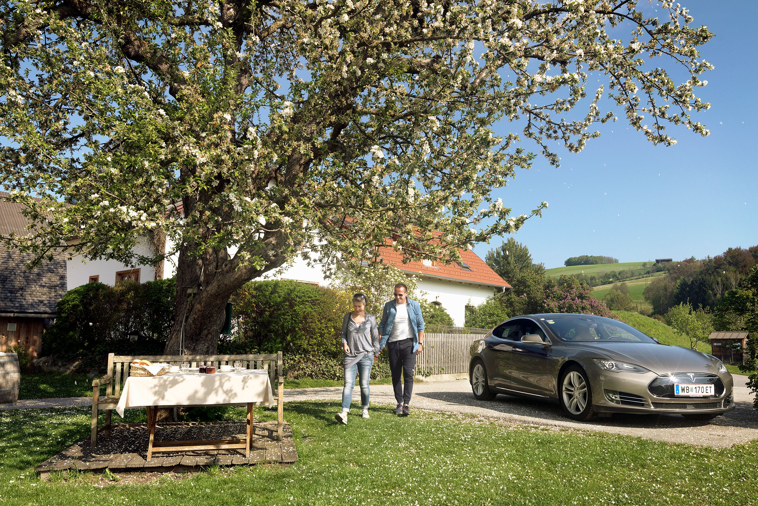 Ein Paar geht neben einem geparkten Elektroauto unter einem blühenden Baum in einem ländlichen Garten.