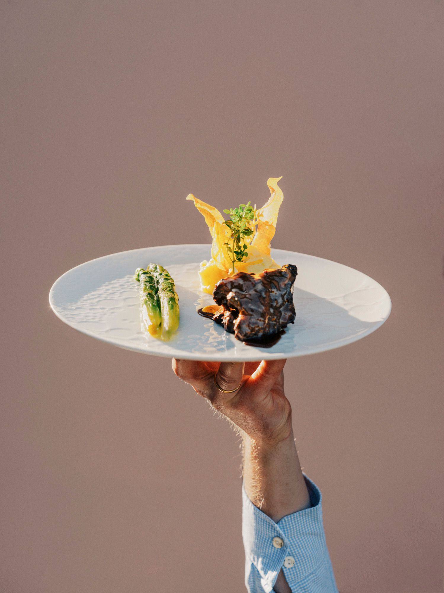 Ein Teller mit Angus-Rind, Spargel und dekorativer Beilage wird von einer Hand hochgehalten.