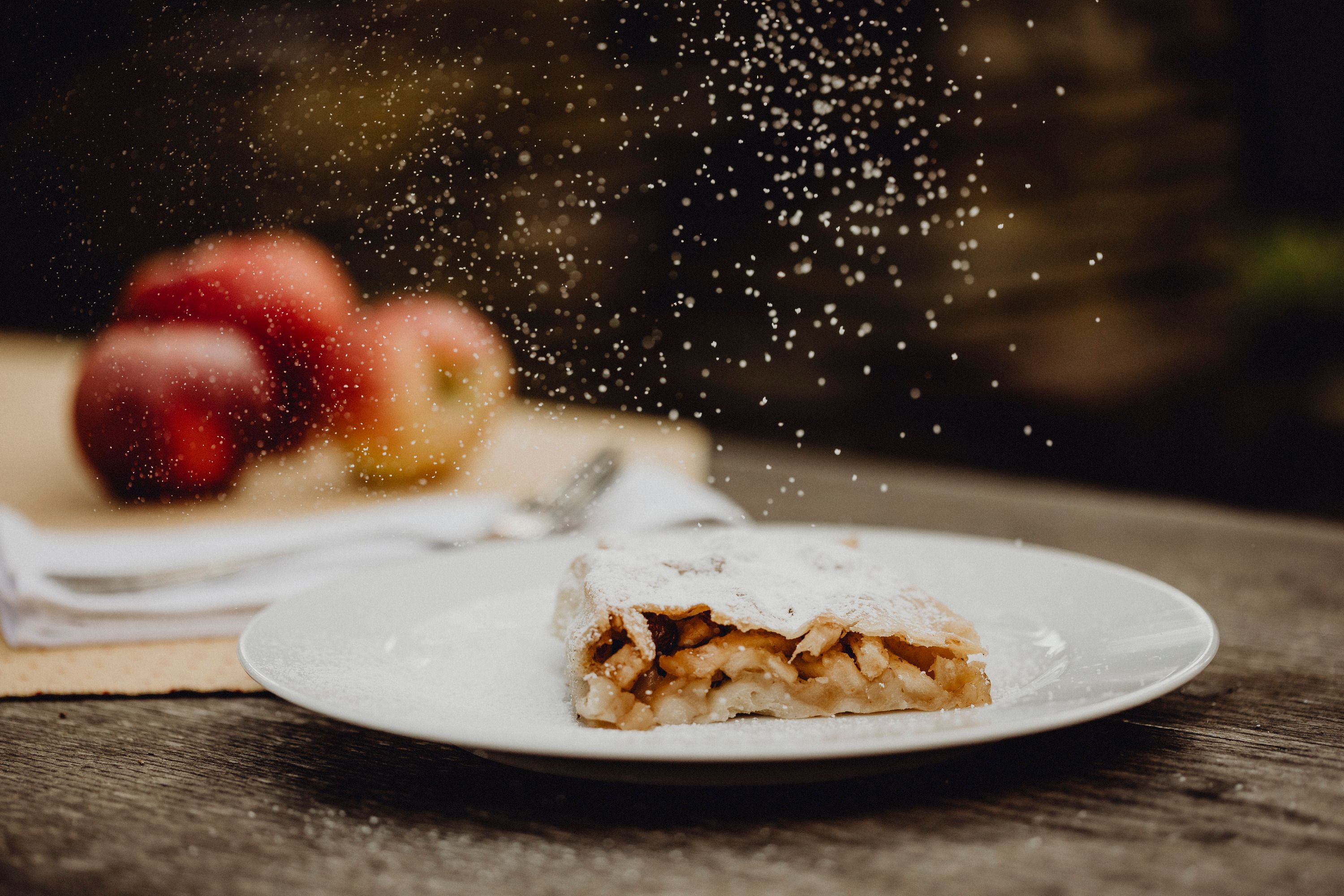 Apfelstrudel mit Puderzucker auf Teller, Äpfel im Hintergrund.