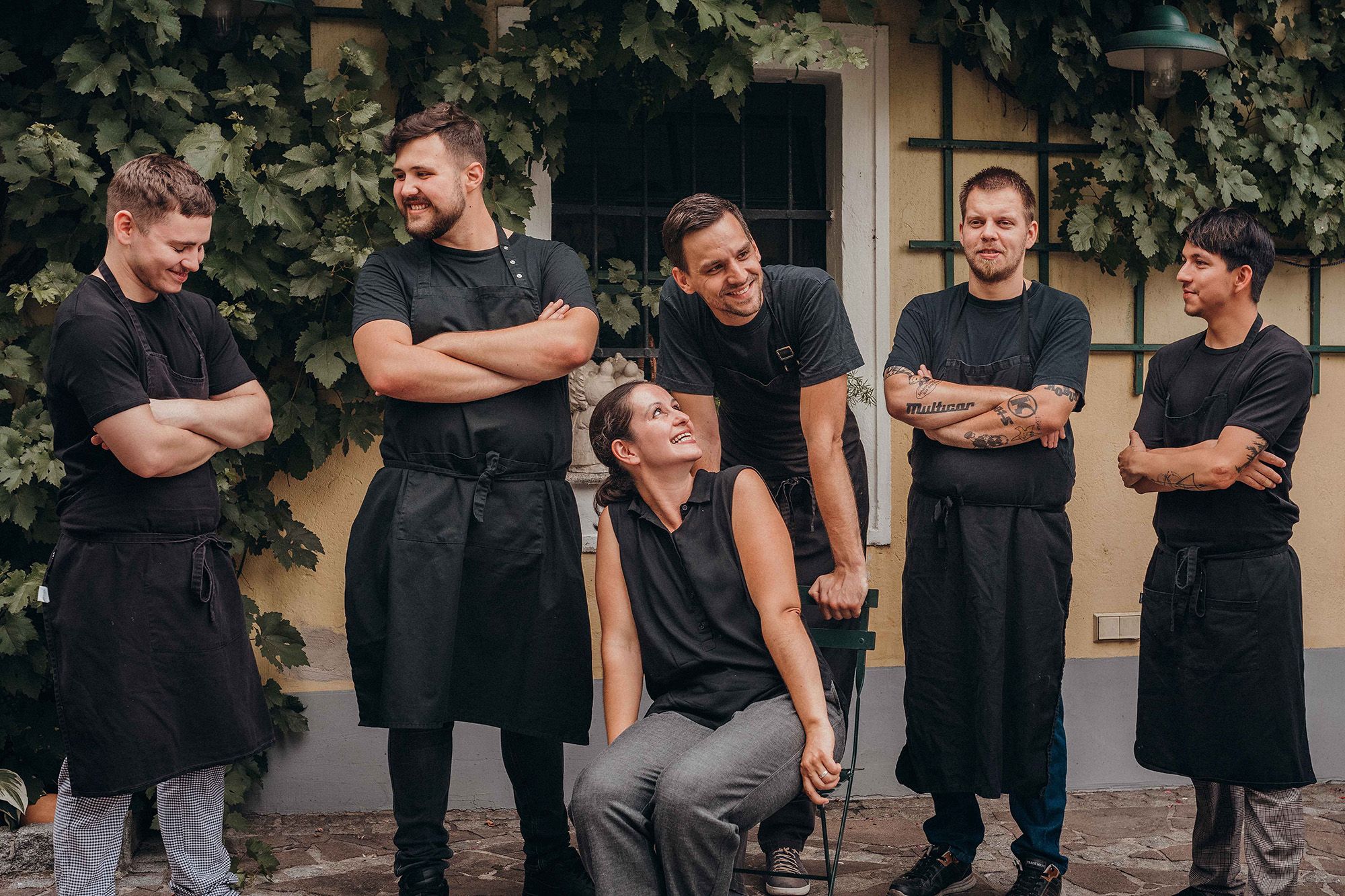 Eine Gruppe von sechs Personen in schwarzer Arbeitskleidung steht und sitzt lächelnd vor einer mit Weinreben bewachsenen Wand.