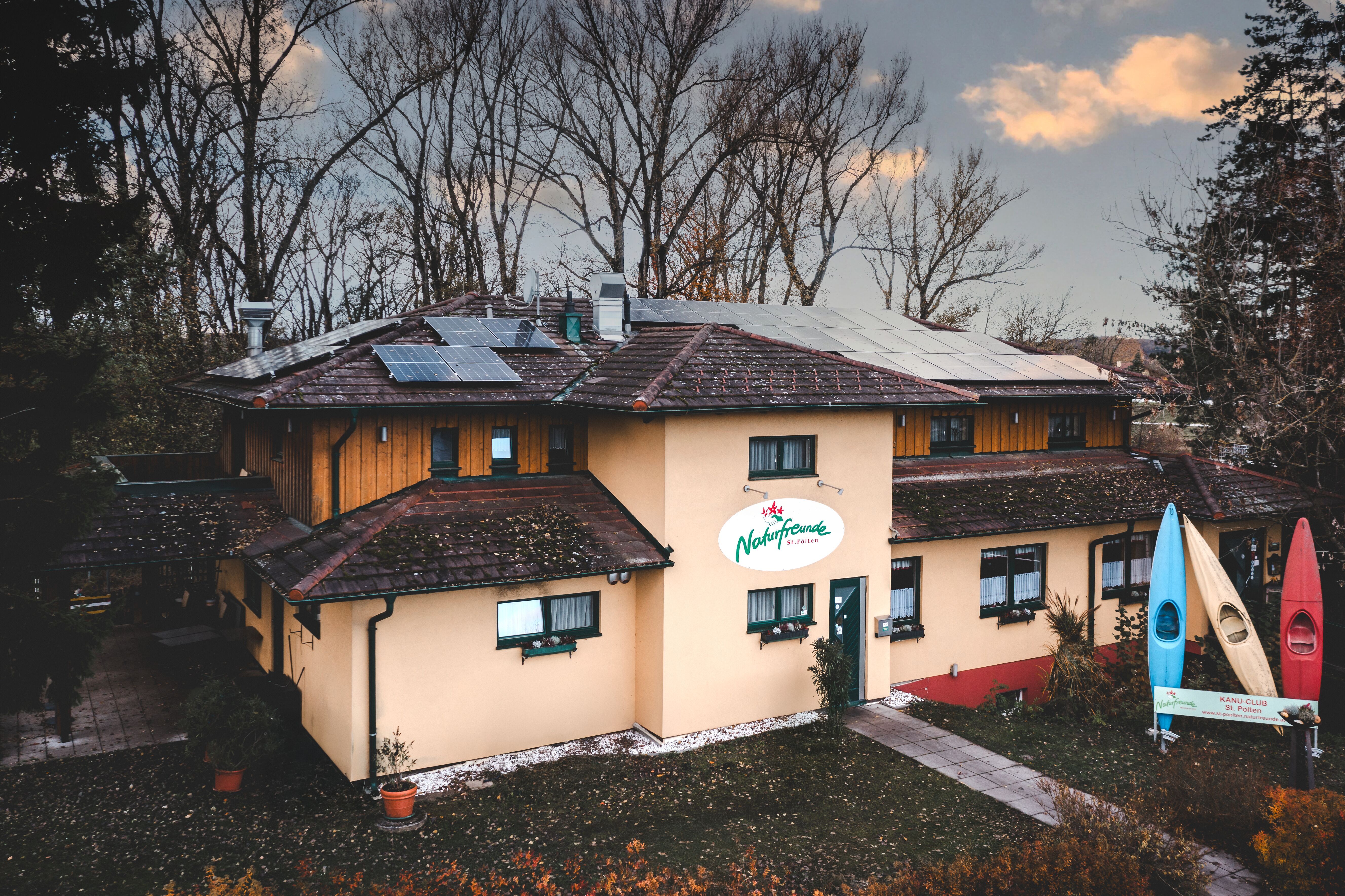 Ein Gebäude mit Solarpanelen auf dem Dach und einem Schild mit der Aufschrift 'Naturfreunde'. Vor dem Gebäude stehen drei bunte Kanus.