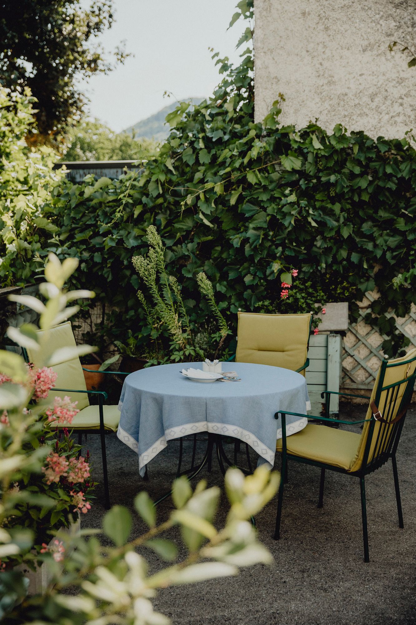 A round table with a blue tablecloth and four chairs in a green garden.