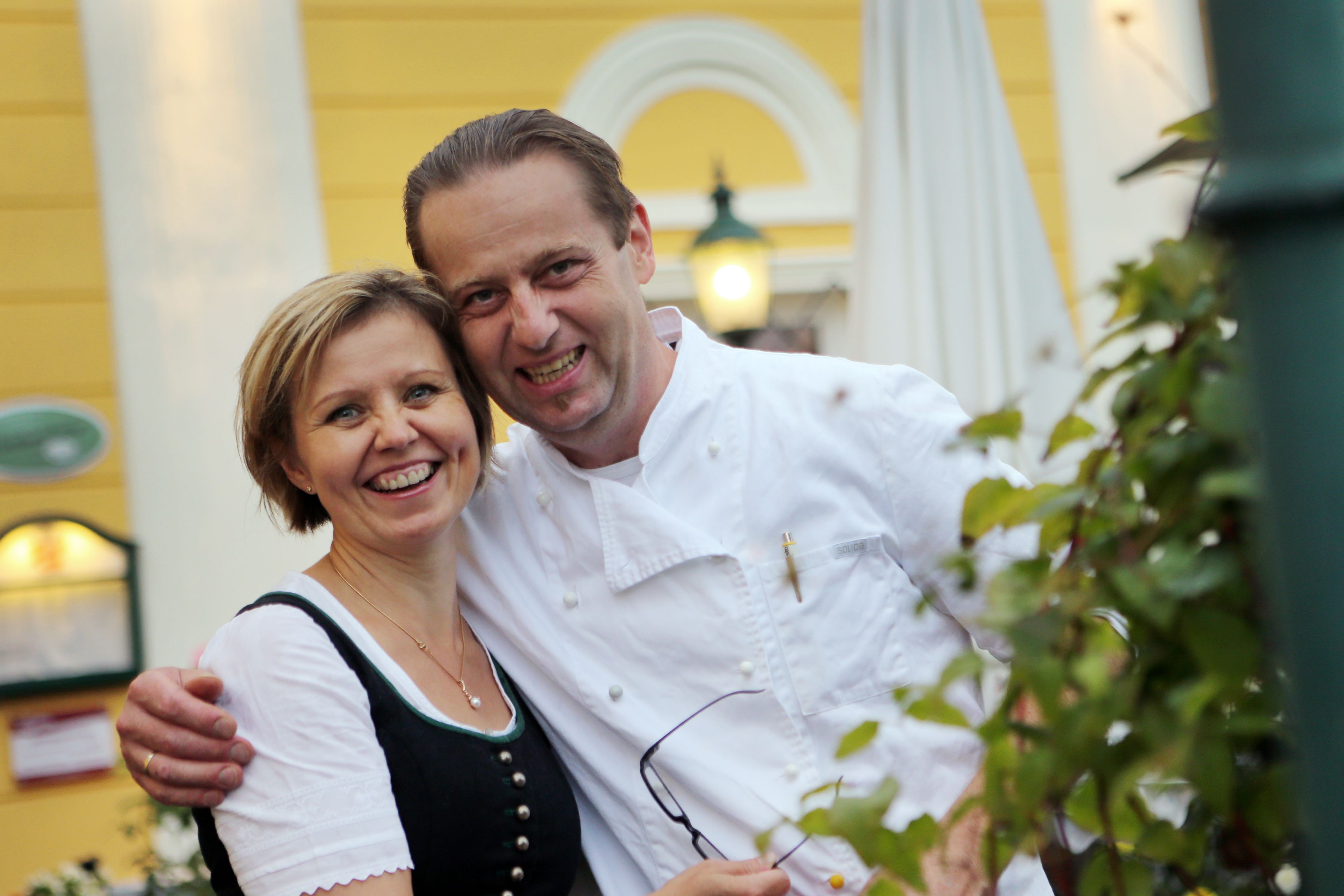 A smiling couple in traditional dress in front of a yellow building.