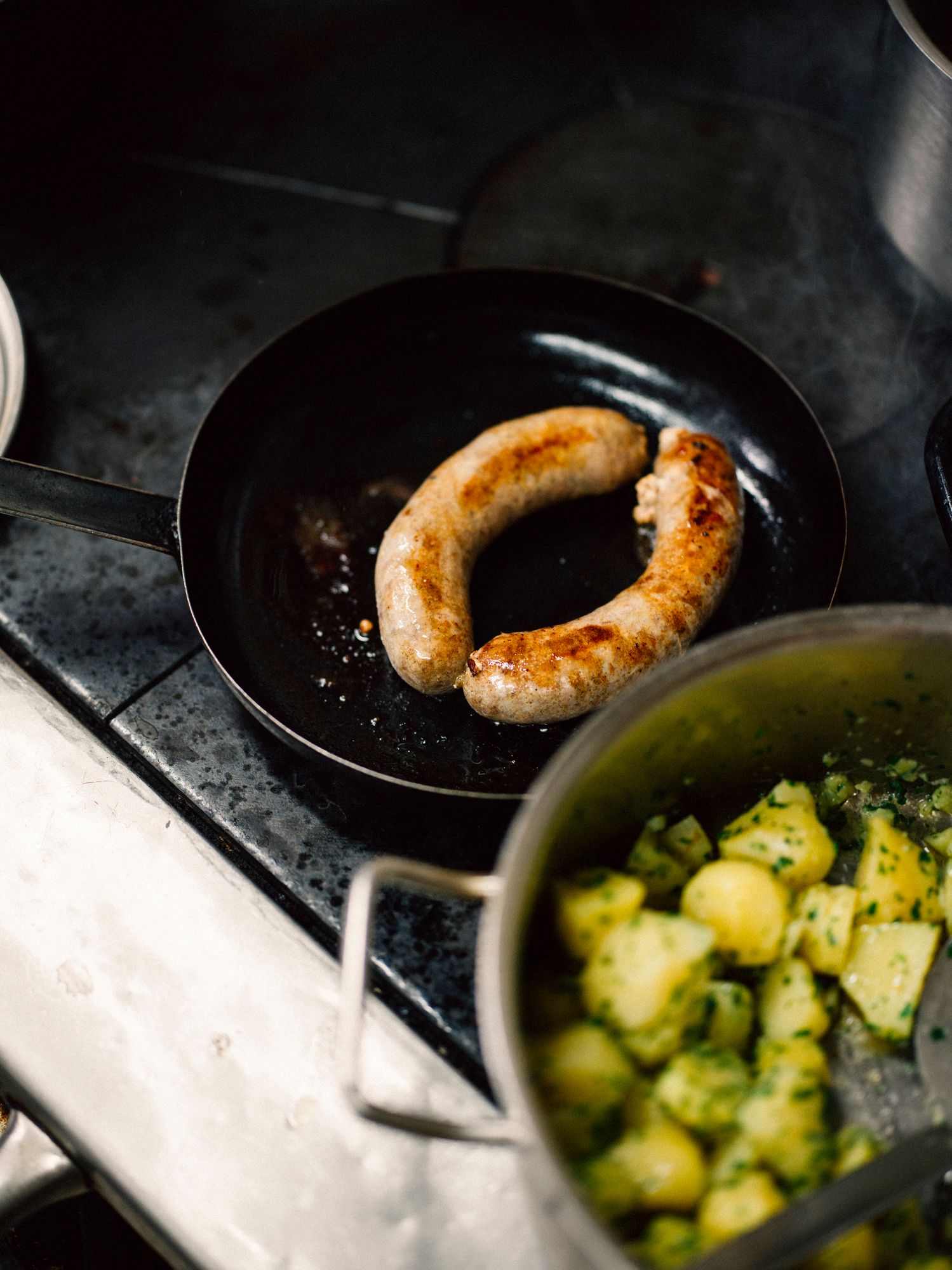 Zwei Bratwürste in einer Pfanne neben einem Topf mit Kartoffeln.