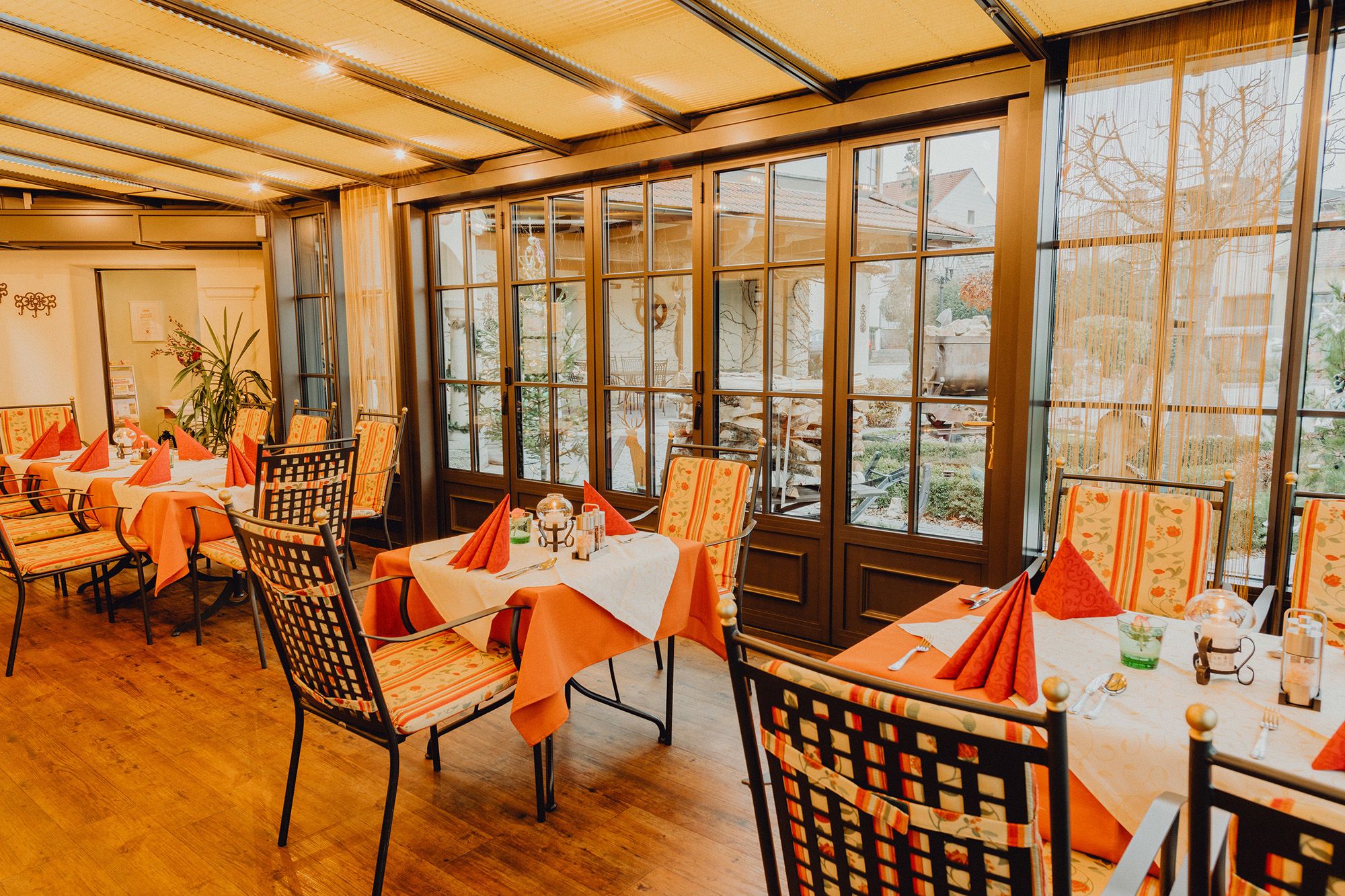 Ein gemütlicher Wintergarten mit gedeckten Tischen und Stühlen, umgeben von großen Fenstern mit Blick nach draußen.