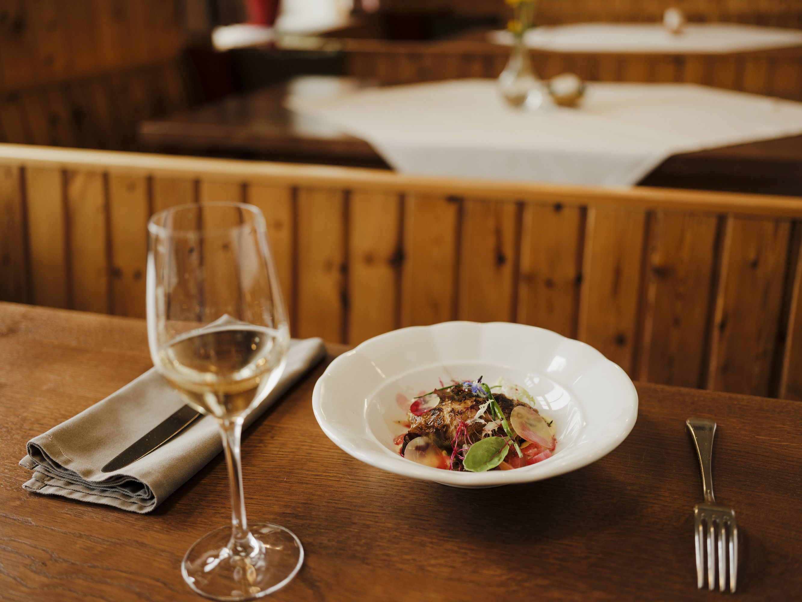 Ein Glas Weißwein und ein Teller mit Gourmetgericht auf einem Holztisch in einem rustikalen Restaurant.