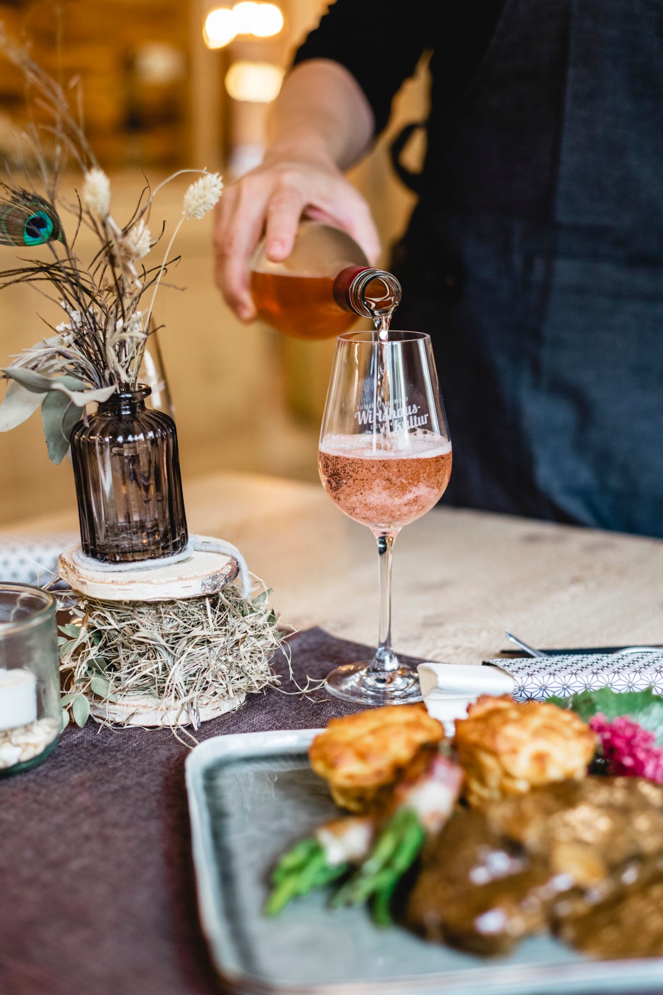 Person schenkt roséfarbenen Wein in ein Glas ein, daneben ein Teller mit Essen und eine Vase mit Trockenblumen.