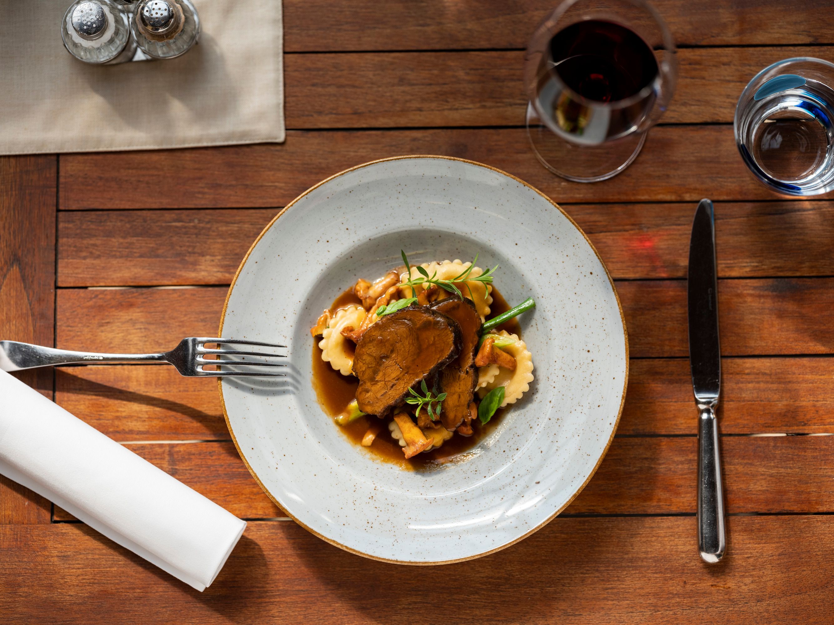 Ein Teller mit geschmorten Rindsbackerl, Ravioli und Gemüse auf einem Holztisch, daneben Besteck und ein Glas Rotwein.