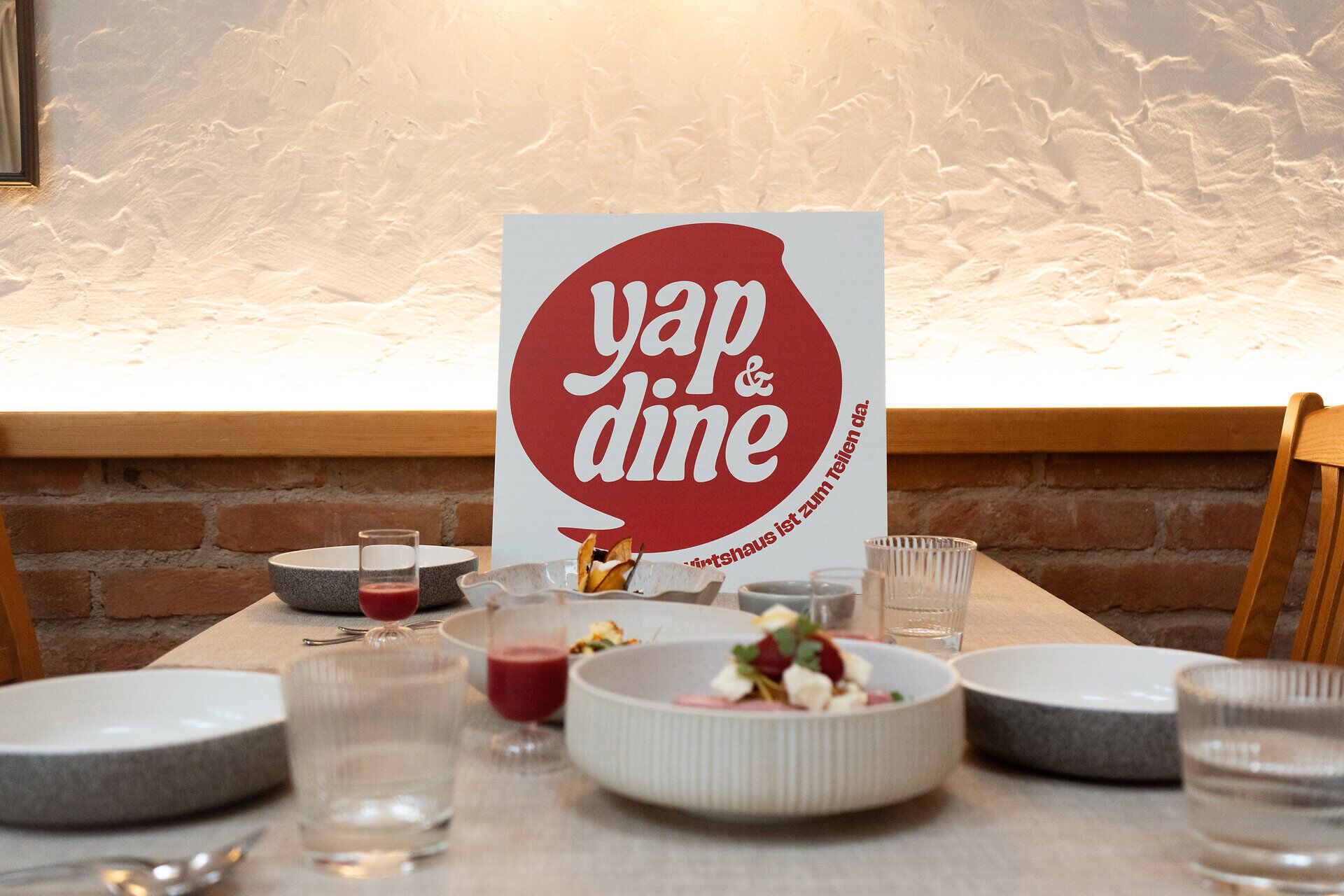 Gedeckter Tisch mit verschiedenen Gerichten und Getränken bei „yap&dine“. Im Hintergrund steht ein Schild mit dem „yap&dine“-Logo und dem Text "Das Wirtshaus ist zum Teilen da.".