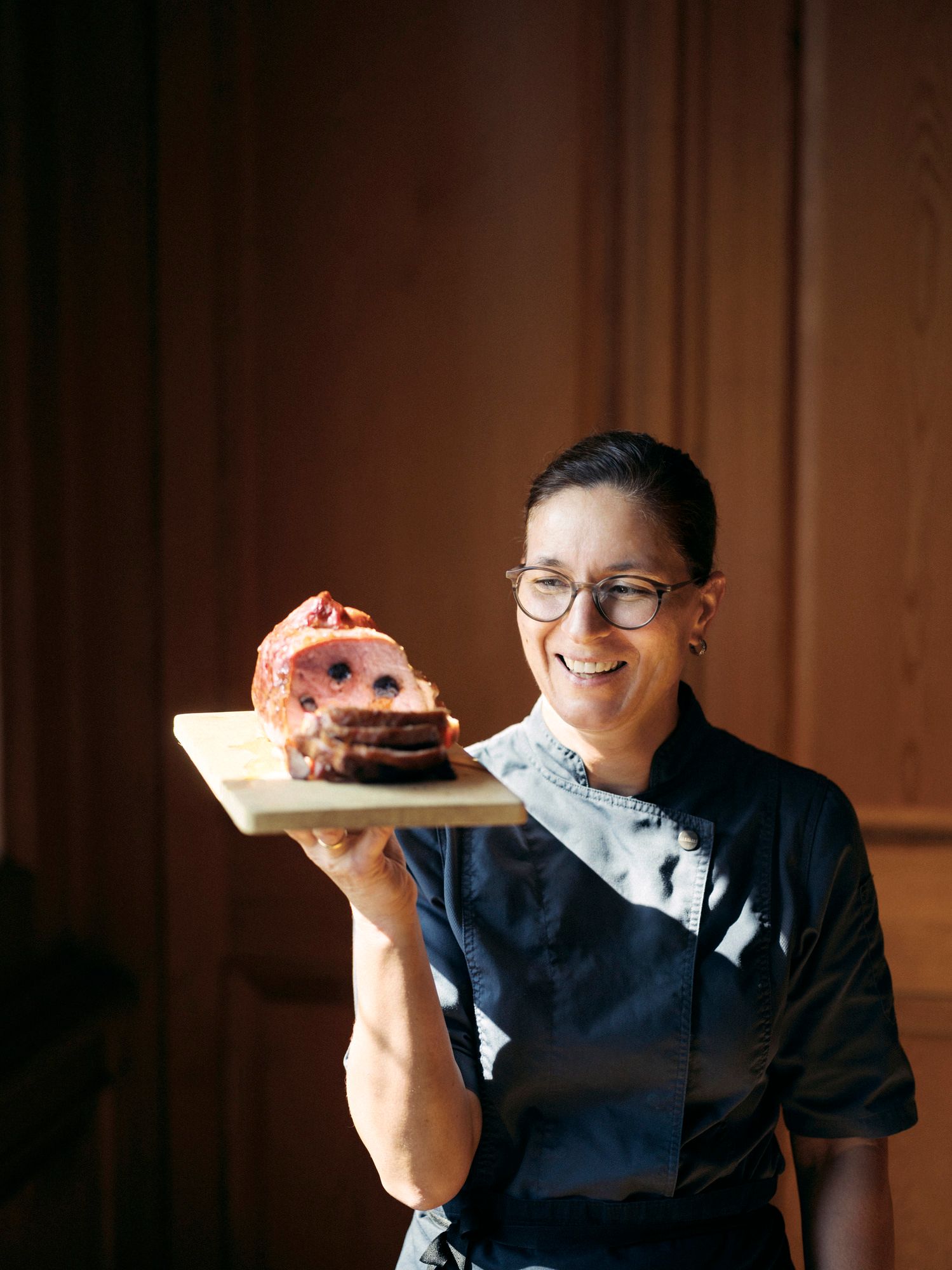 Eine Frau in Kochkleidung hält ein Holzbrett mit einem aufgeschnittenen Fleischgericht.