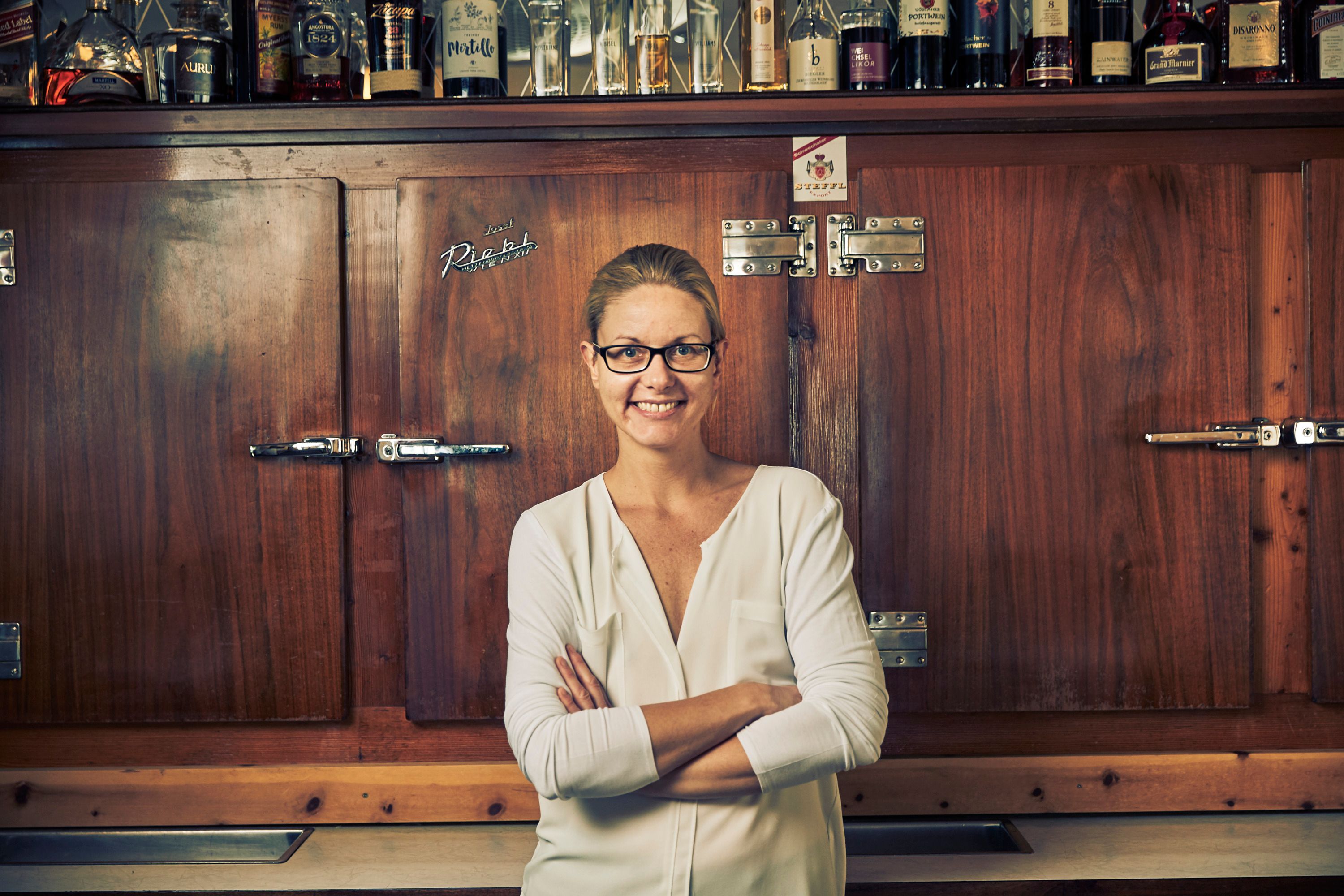 Eine Frau mit Brille steht lächelnd vor einem Holzschrank mit Flaschen darauf.