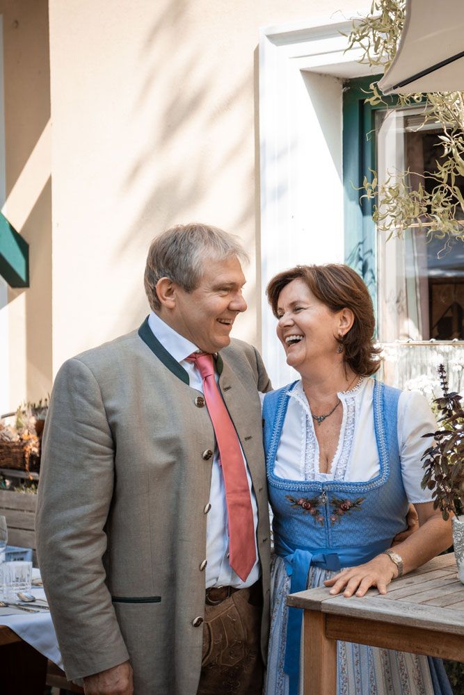 Ein Mann und eine Frau in traditioneller bayerischer Kleidung lachen zusammen vor einem Gebäude.