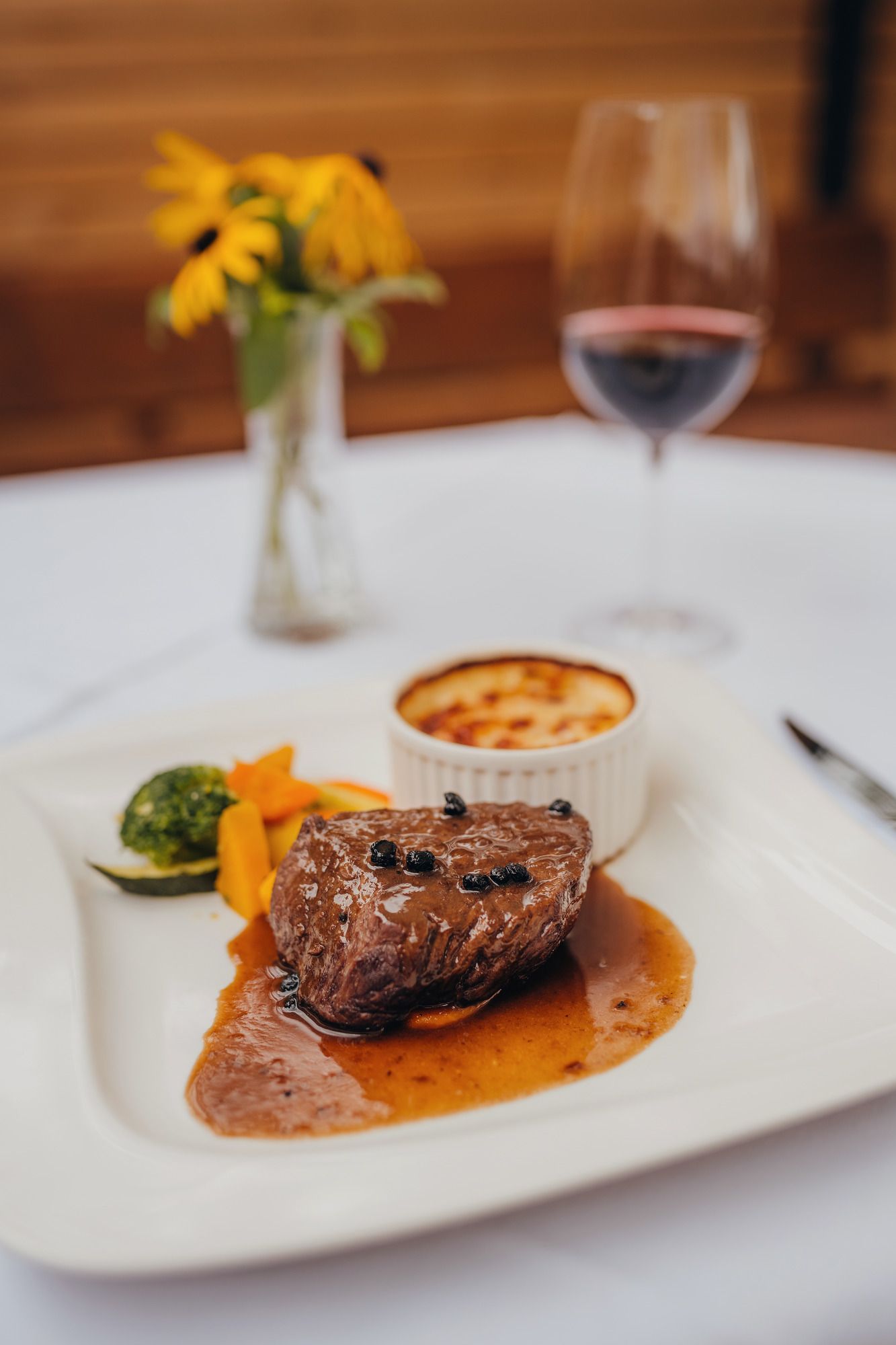 Ein Teller mit Pfeffersteak, Gemüse und Gratin, daneben ein Glas Rotwein und eine Vase mit gelben Blumen.