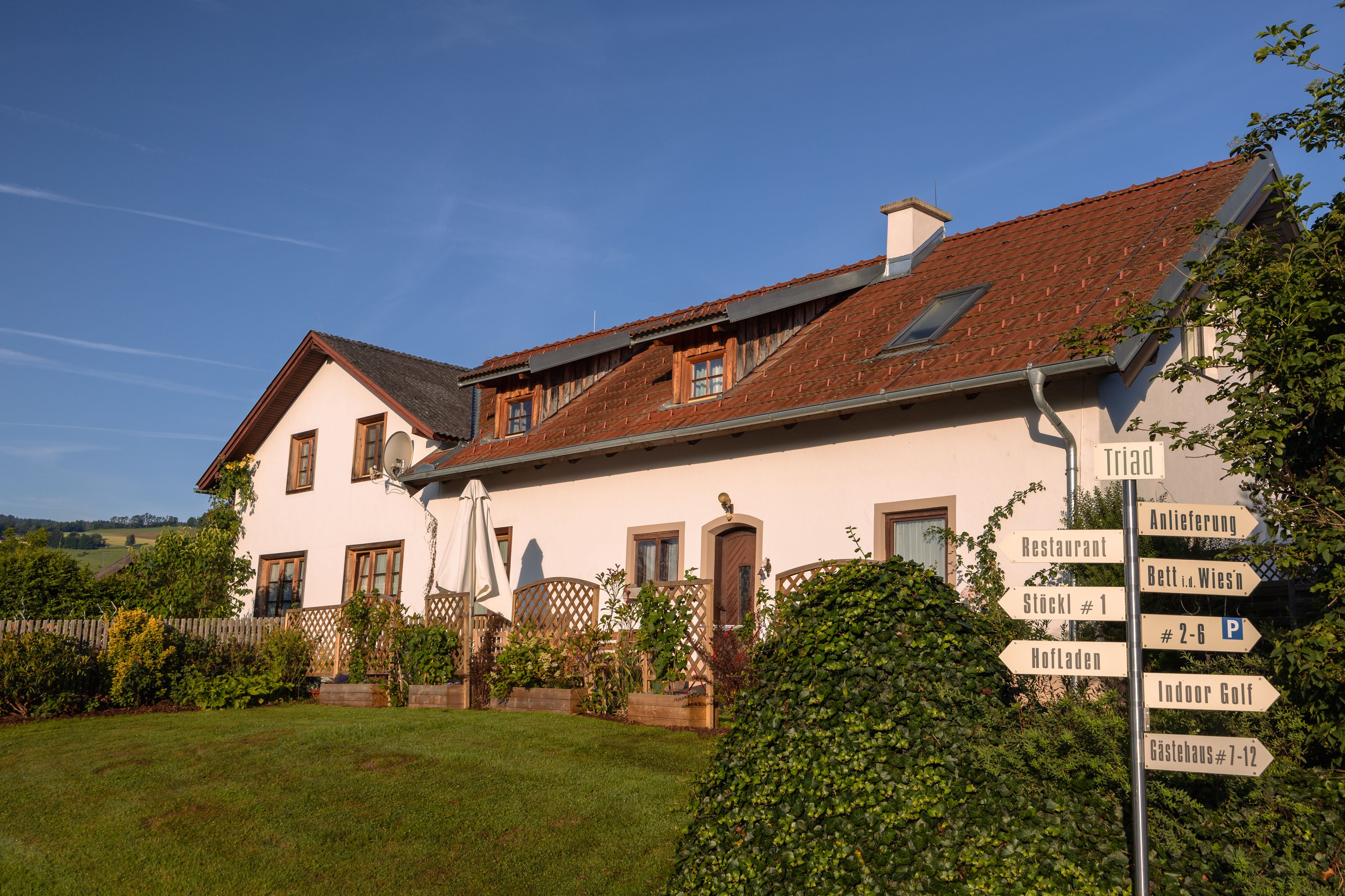 Ein weißes Gebäude mit rotem Dach und mehreren Schildern im Vordergrund, umgeben von grüner Landschaft.