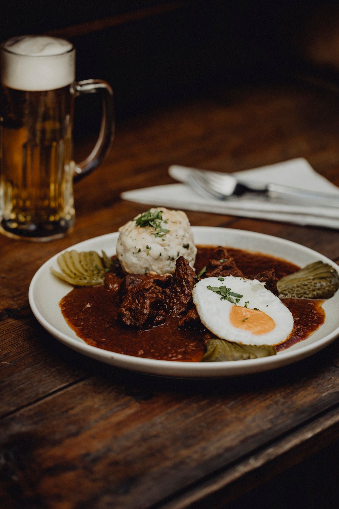Ein Teller mit Gulasch, Spiegelei, Knödel und Gurken auf einem Holztisch, daneben ein Bierkrug.