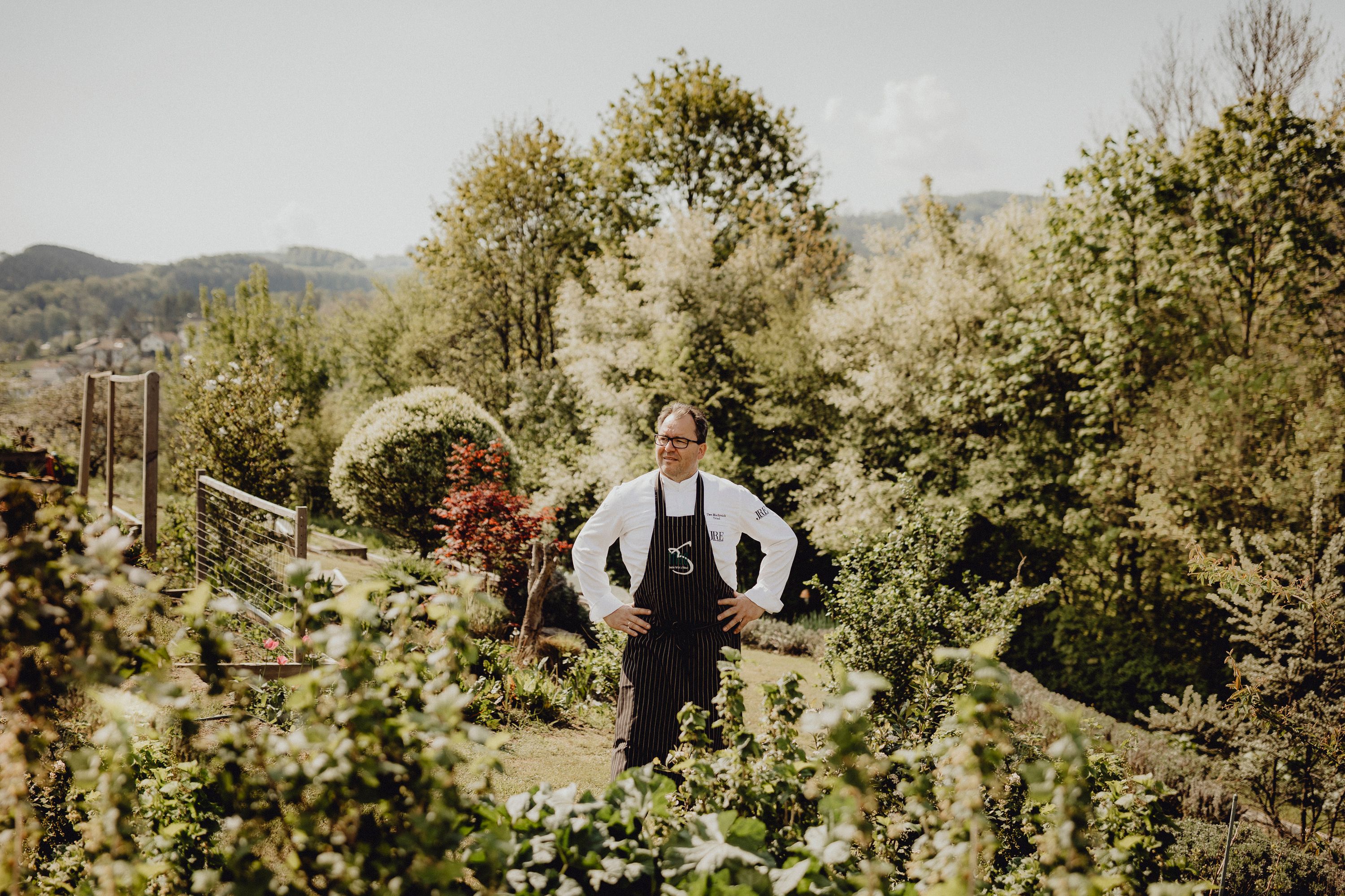 Ein Koch steht in einem Garten mit Bäumen im Hintergrund.