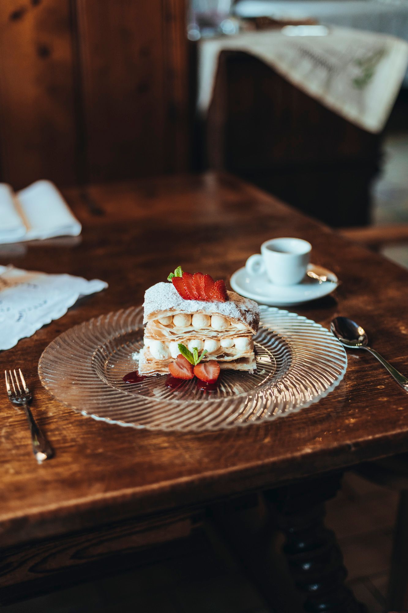 Glasteller mit Cremeschnitte, Erdbeeren und Minze, steht auf einem Holztisch neben einem Espresso.