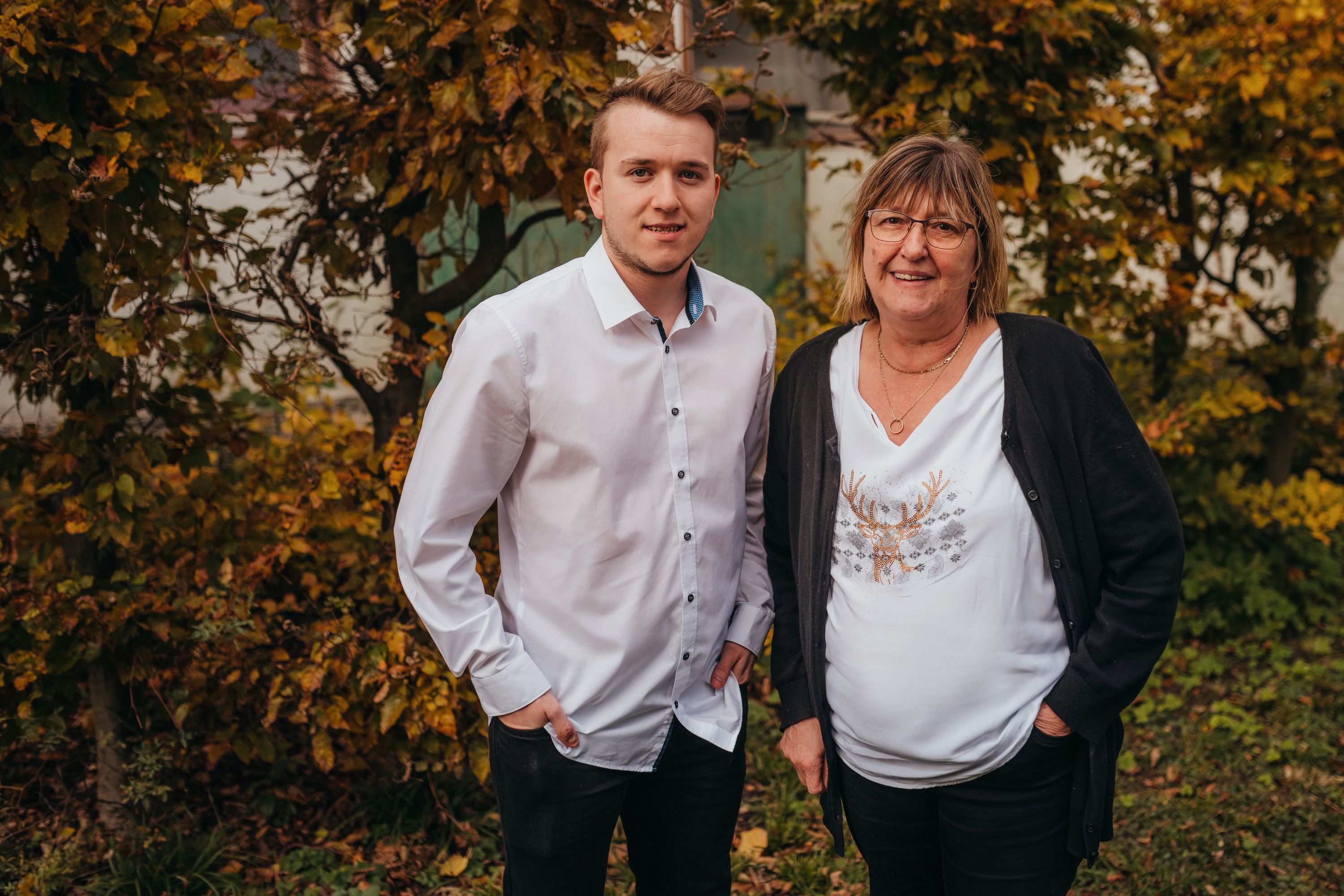 Ein junger Mann und eine ältere Frau stehen lächelnd vor herbstlichen Bäumen.
