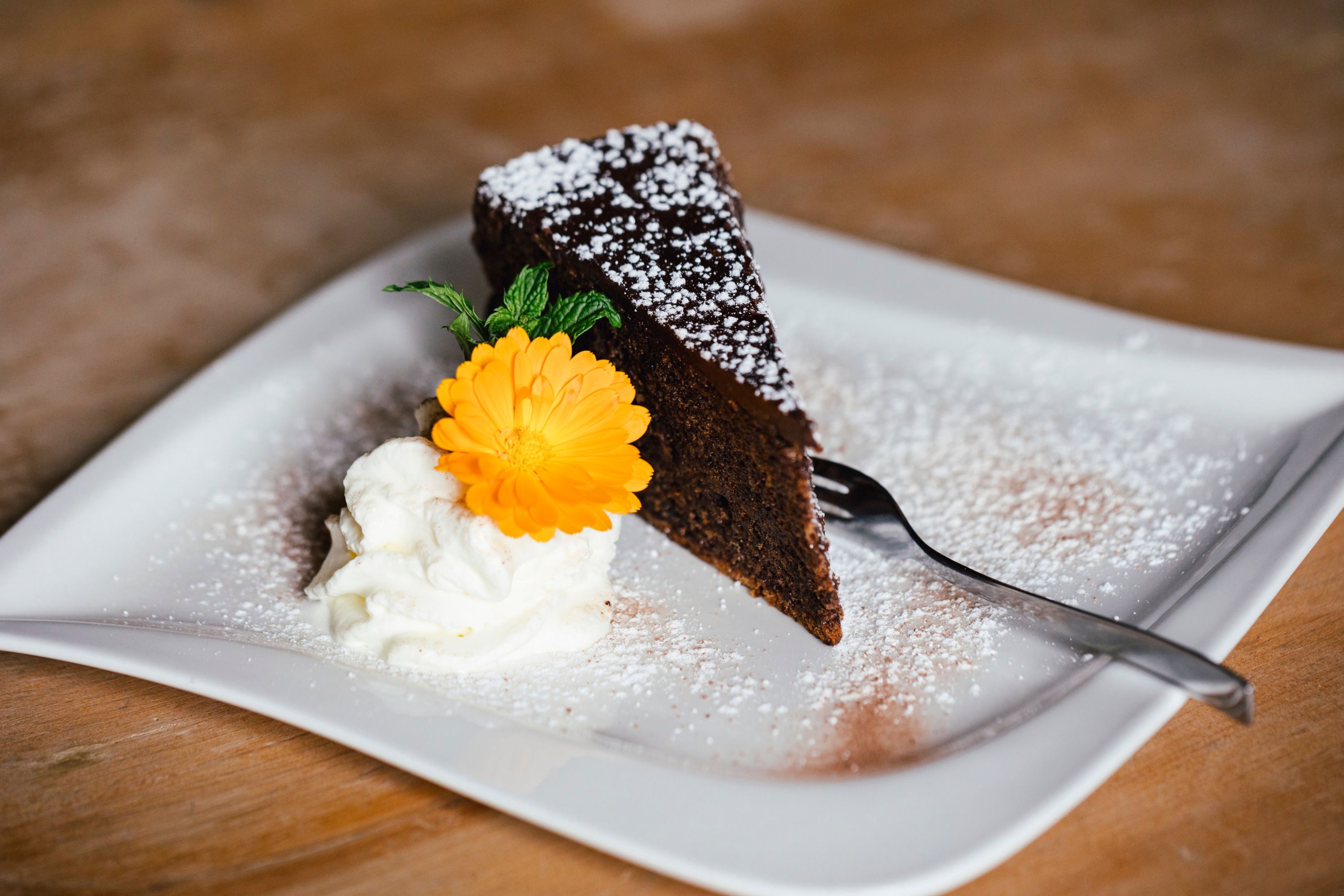 Ein Stück Schokoladenkuchen mit Puderzucker, Sahne und einer gelben Blume auf einem Teller.