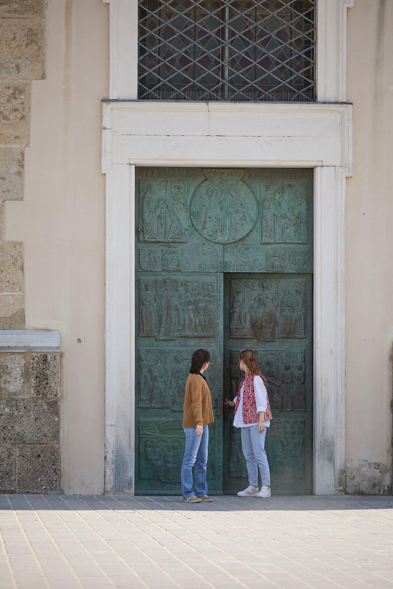 Zwei Frauen stehen fasziniert vor einer kunstvoll gestalteten Tür, die mit historischen Reliefs verziert ist. Die warme Sonne taucht die Szene in ein sanftes Licht und schafft eine einladende Atmosphäre, die zum Verweilen einlädt. Umgeben von der beeindruckenden Architektur des Regierungsviertels, spürt man die Verbindung von Geschichte und Gegenwart.
