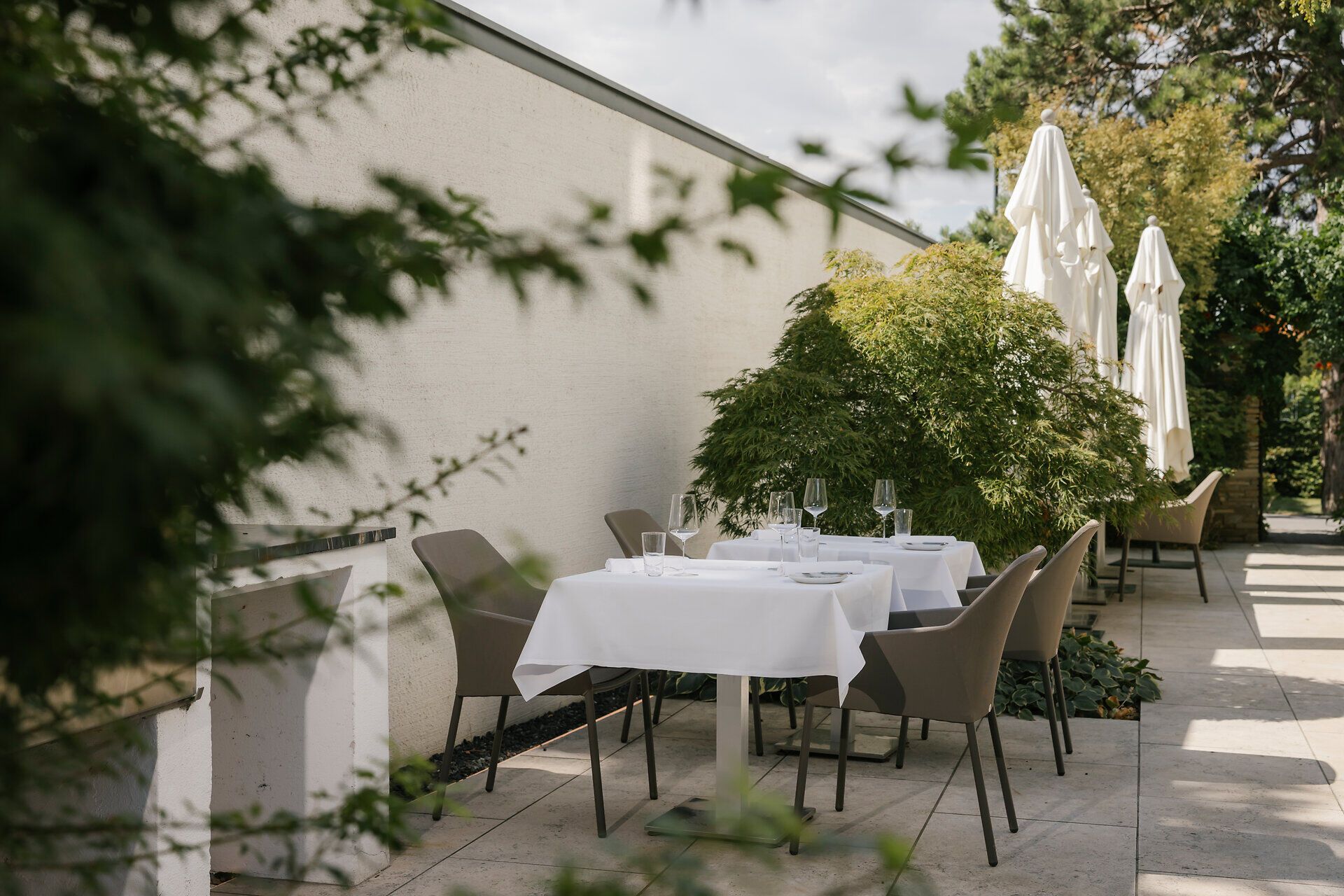 Gastgarten mit weiß gedeckten Tischen, grauen Stühlen und Weingläsern. Zwischen grünen Sträuchern stehen mehrere geschlossene weiße Sonnenschirme.