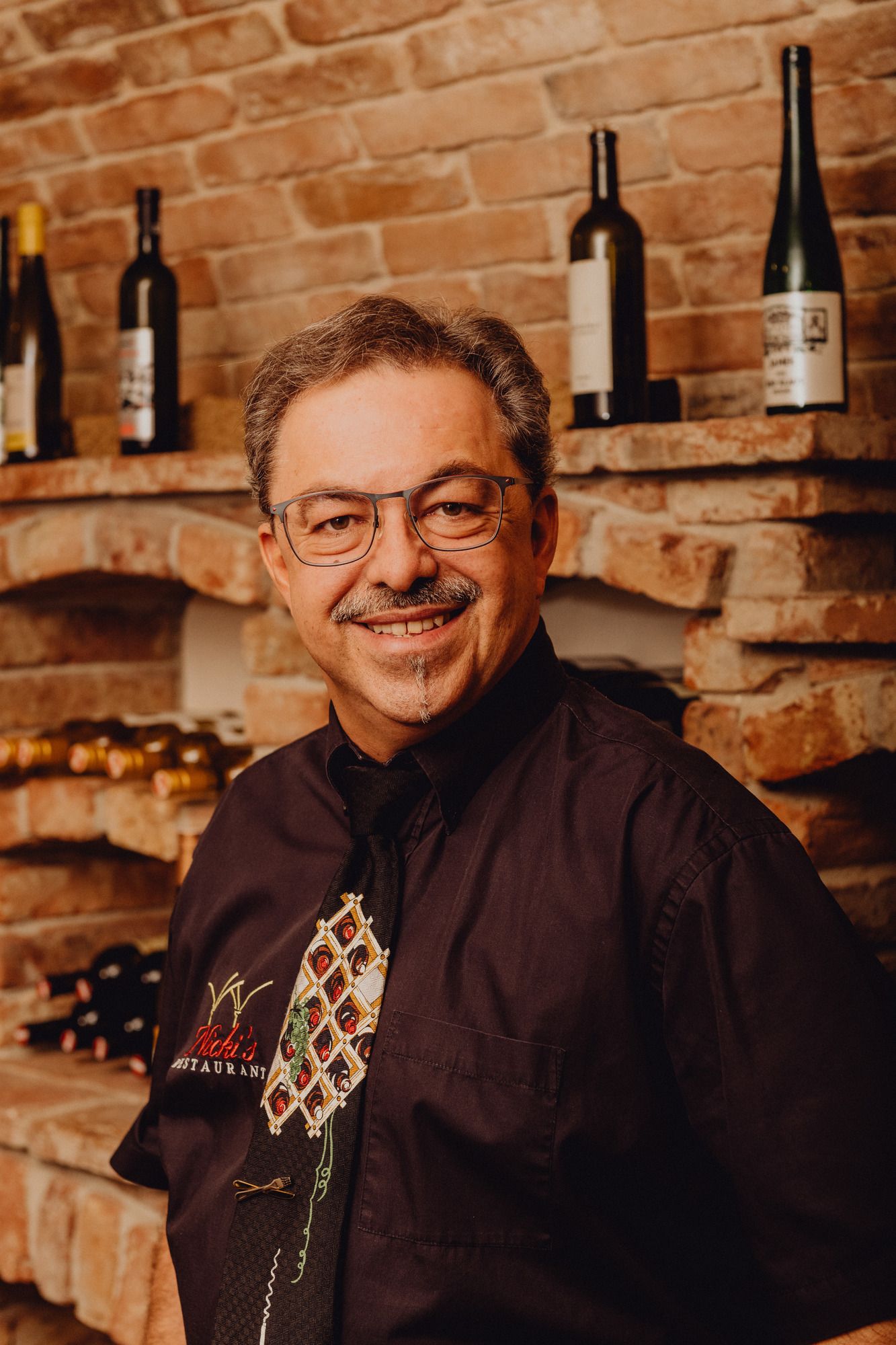 Ein Mann mit Brille und Schnurrbart steht vor einem Weinregal aus Backstein.