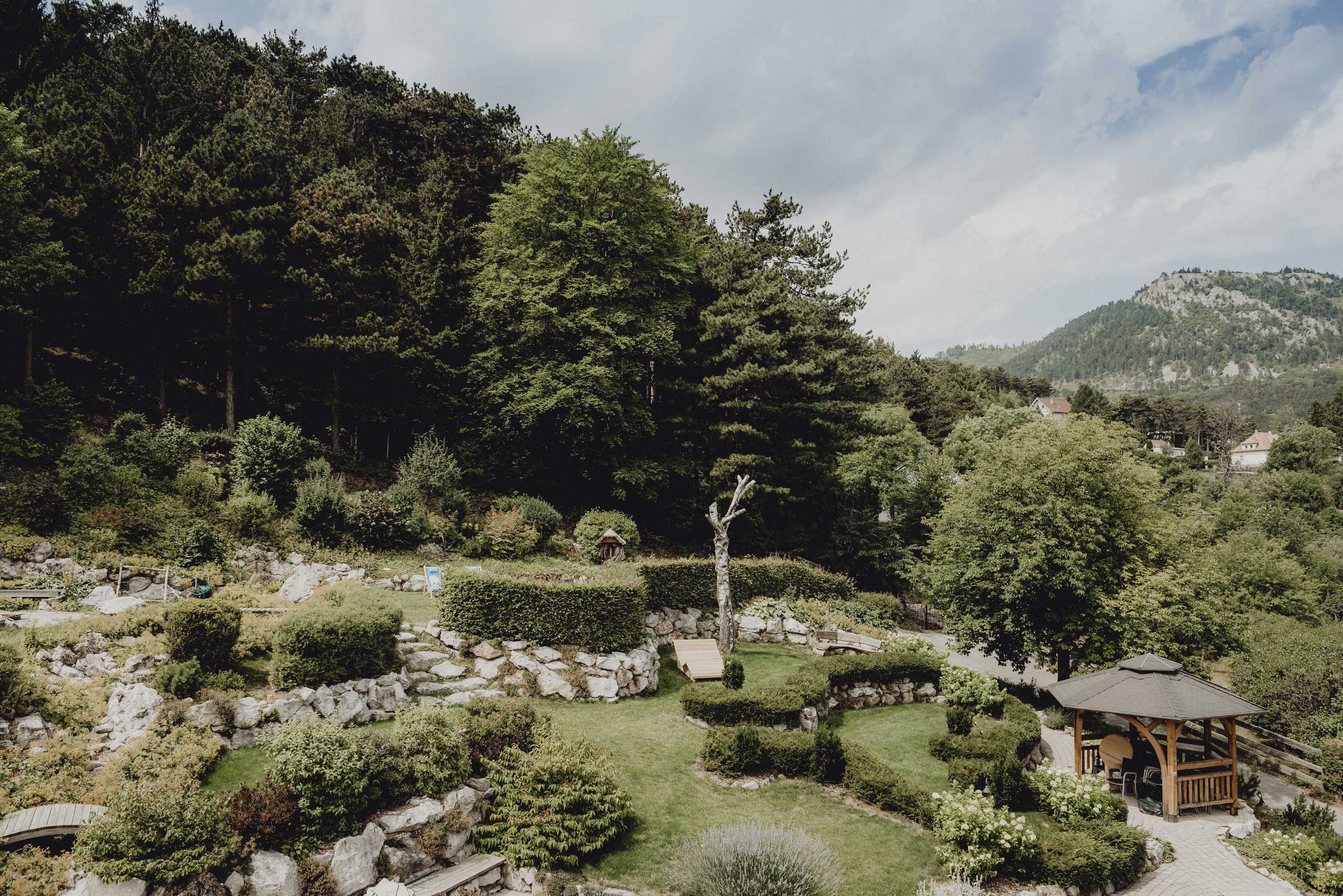 Ein Alpengarten mit Bäumen, Sträuchern und einem Pavillon vor einer bewaldeten Berglandschaft.