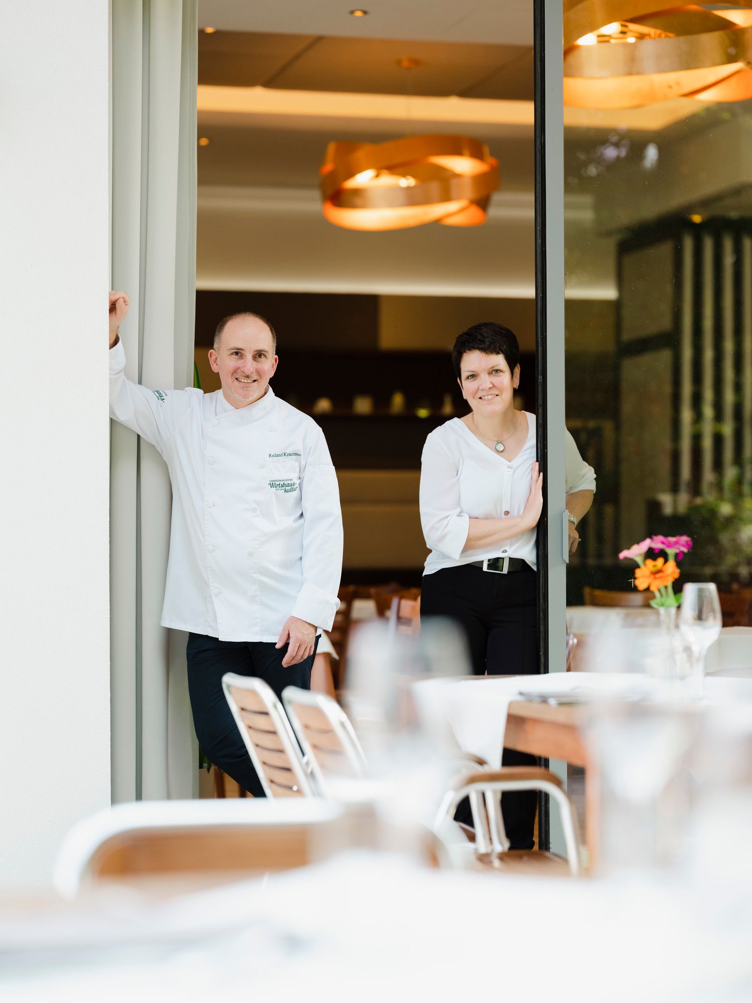 Zwei Personen stehen in einem Restaurant am Eingang, eine in Kochkleidung, die andere in weißer Bluse.