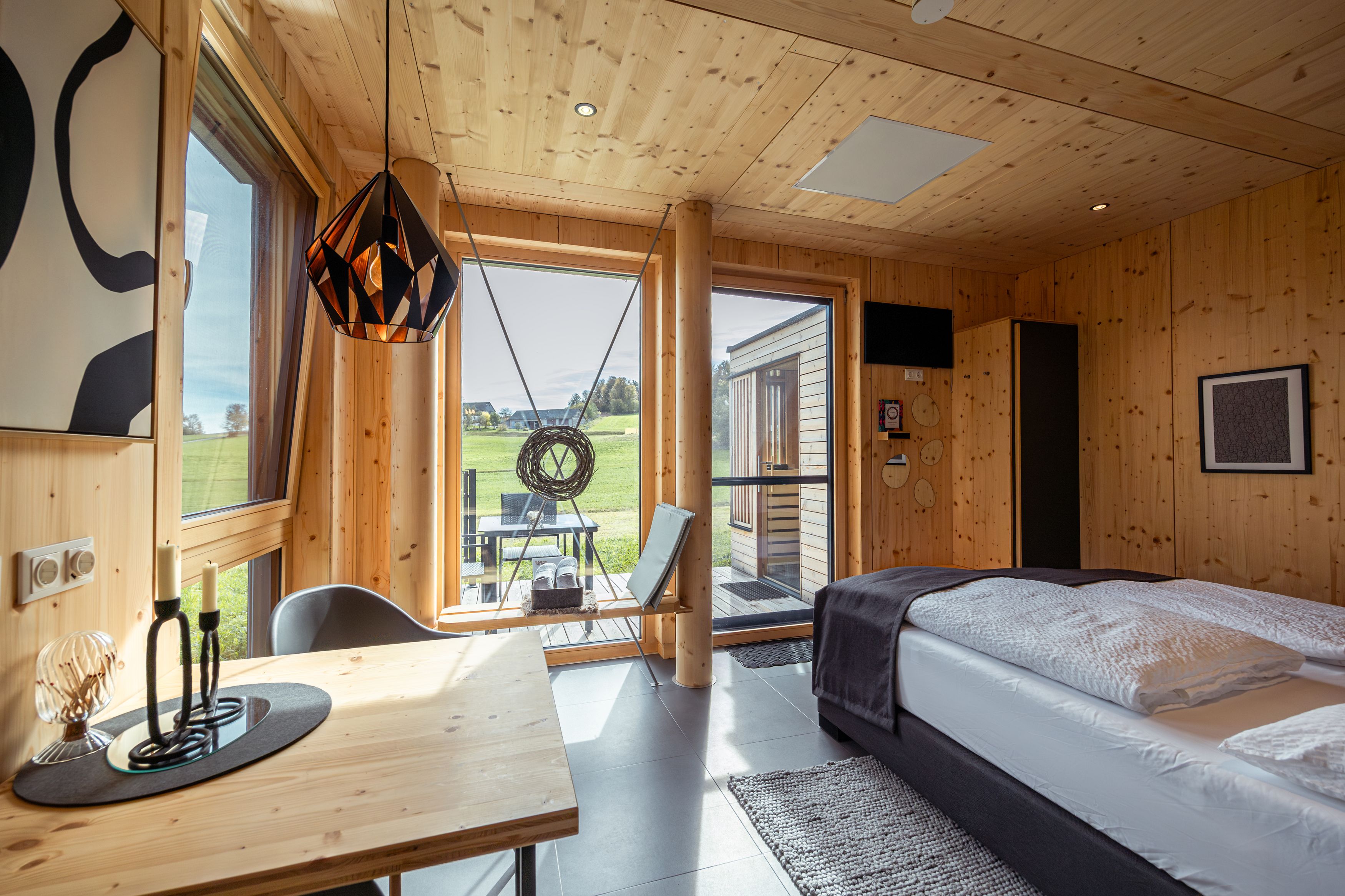 Innenansicht eines modernen Holzhauses mit Bett, Tisch und großen Fenstern mit Blick ins Freie.