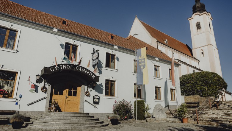 Ein Gasthof mit der Aufschrift 'Gasthof Gamerith' neben einer Kirche mit Turm unter blauem Himmel.