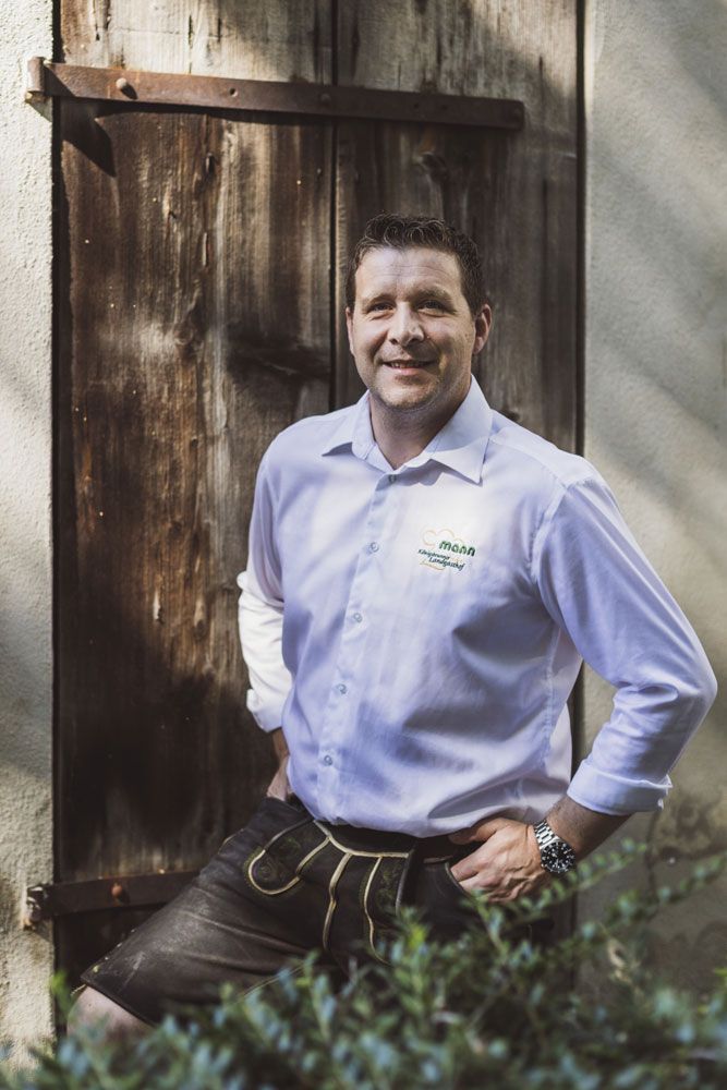 Mann in Tracht vor Holztür, trägt Hemd mit Logo, lächelt in die Kamera.