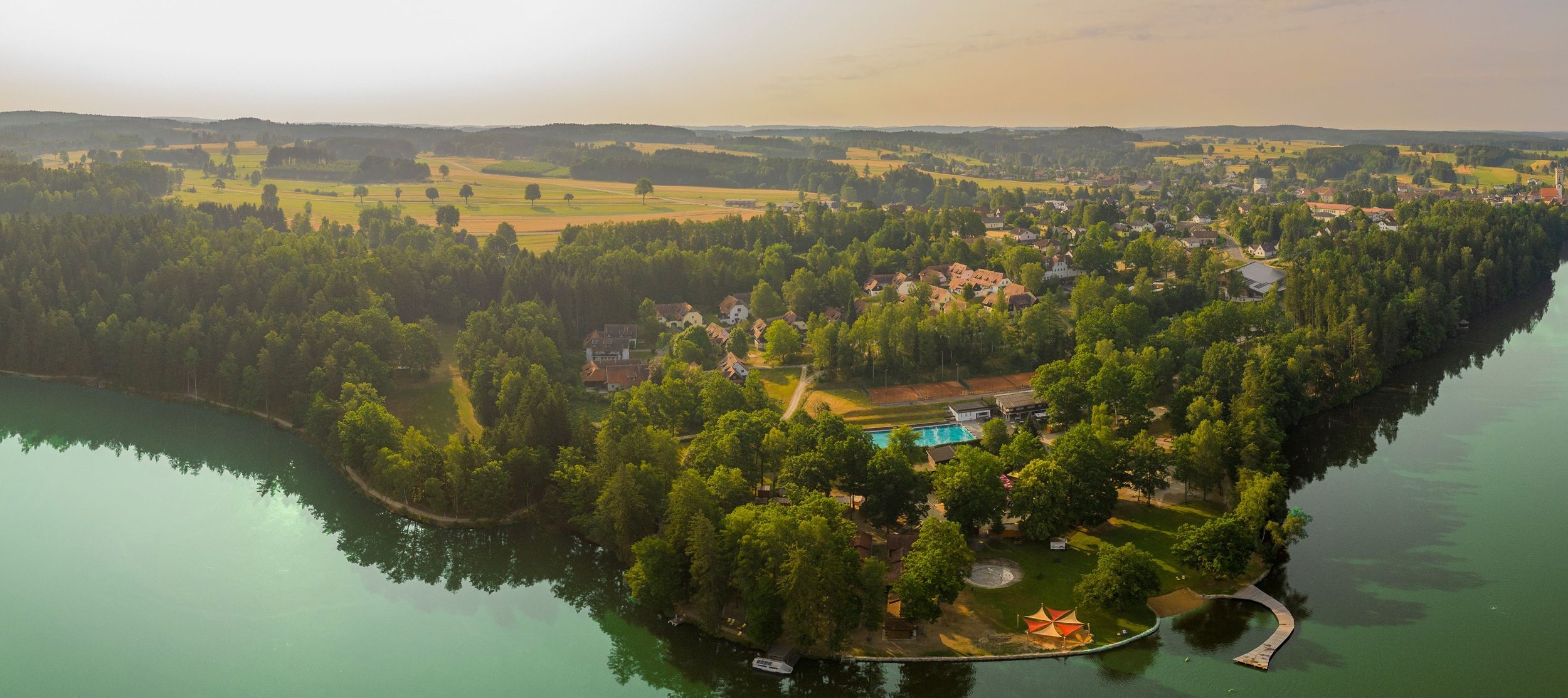 Luftaufnahme eines Sees mit umliegendem Wald und Gebäuden.