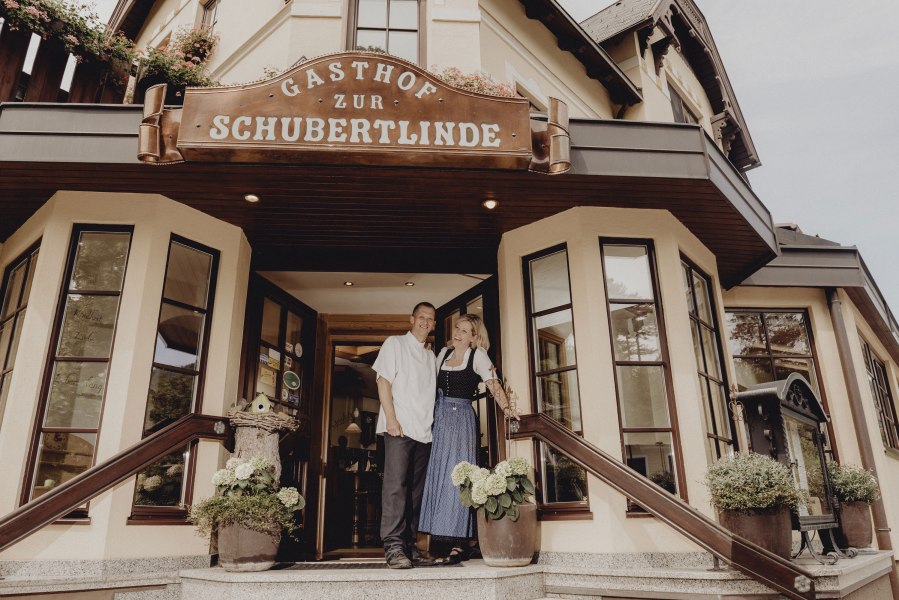 Eingang des Gasthofs 'Zur Schubertlinde' mit zwei Personen in traditioneller Kleidung.