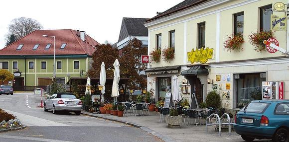 Au&szlig;enansicht von einem Gasthaus an einem Belebten Platz mit B&auml;umen, Sonnenschirmen, Sesseln uvm.