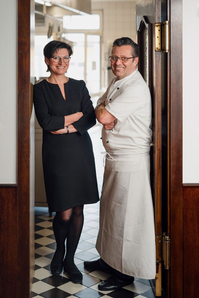 Ein Mann in Kochkleidung und eine Frau in schwarzem Kleid stehen lächelnd in einer Türöffnung.