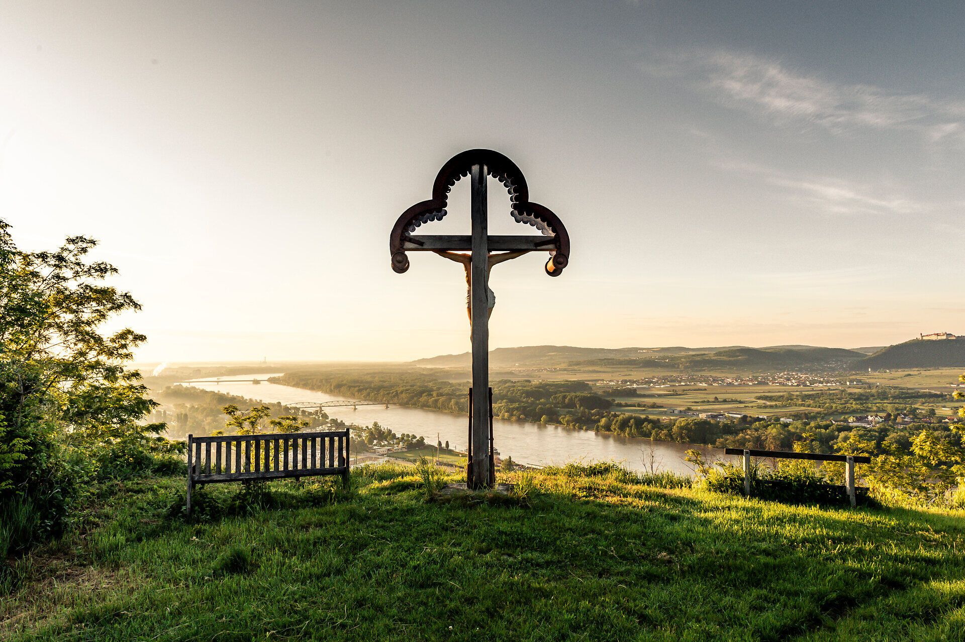 Ein majestätisches Kreuz erhebt sich über die sanften Hügel, während die Donau friedlich im Sonnenlicht glitzert. Die frische Frühlingsluft erfüllt die Umgebung mit einem Gefühl von Freiheit und Abenteuer, ideal für einen unvergesslichen Ausblick. Hier, wo Natur und Spiritualität aufeinandertreffen, lädt die Landschaft dazu ein, innezuhalten und die Schönheit der Region zu genießen.