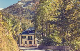 Ein gelbes Geb&auml;ude, umgeben von B&auml;umen, vor einem bewaldeten Berg mit Schnee auf der Spitze.