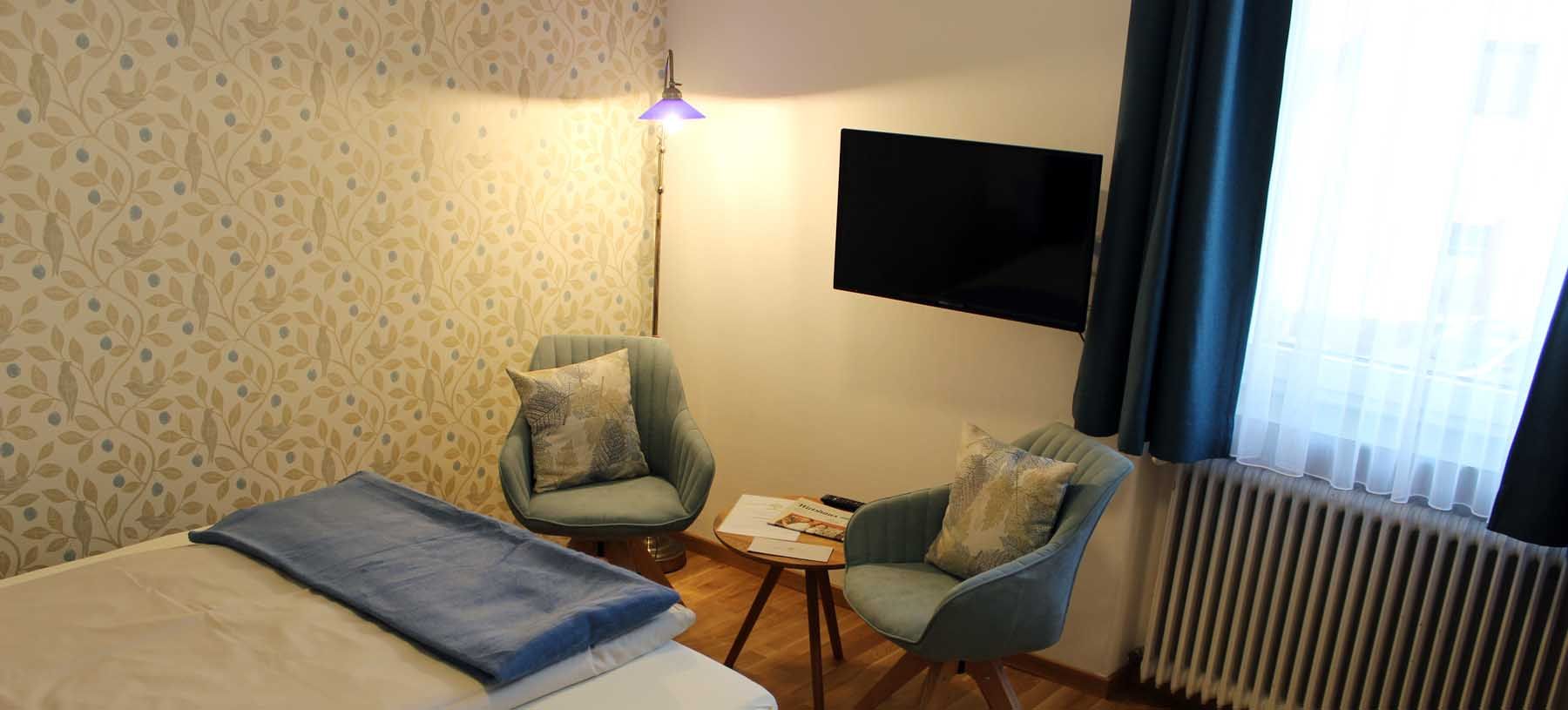 Zimmer mit Bett, Tisch mit zwei Stühlen, Fernseher, Fenster mit blauen Vorhängen und Stehlampe.