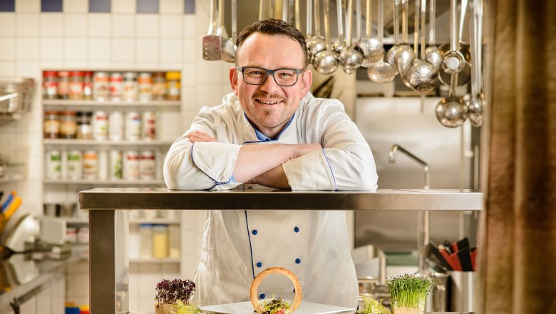 Ein Koch in weißer Uniform lehnt sich auf eine Edelstahltheke in einer Küche. Vor ihm steht ein Teller mit dekorativem Essen.