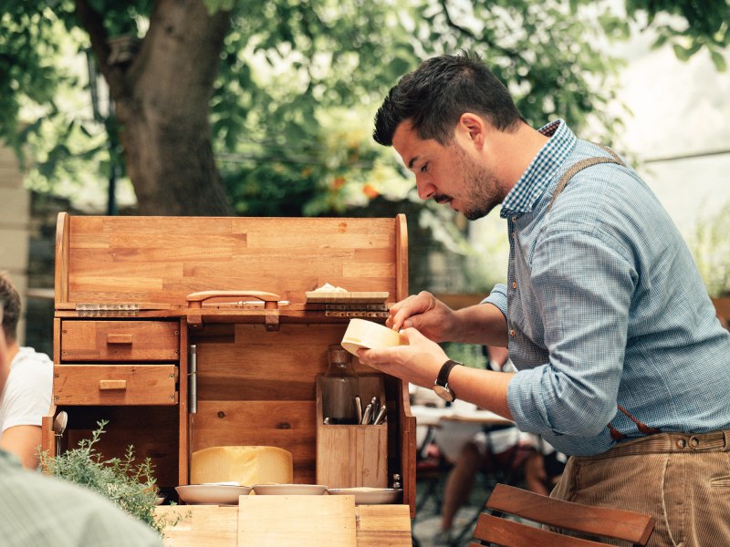 Ein Mann in einem blauen Hemd schneidet K&auml;se an einem Holztisch im Freien.