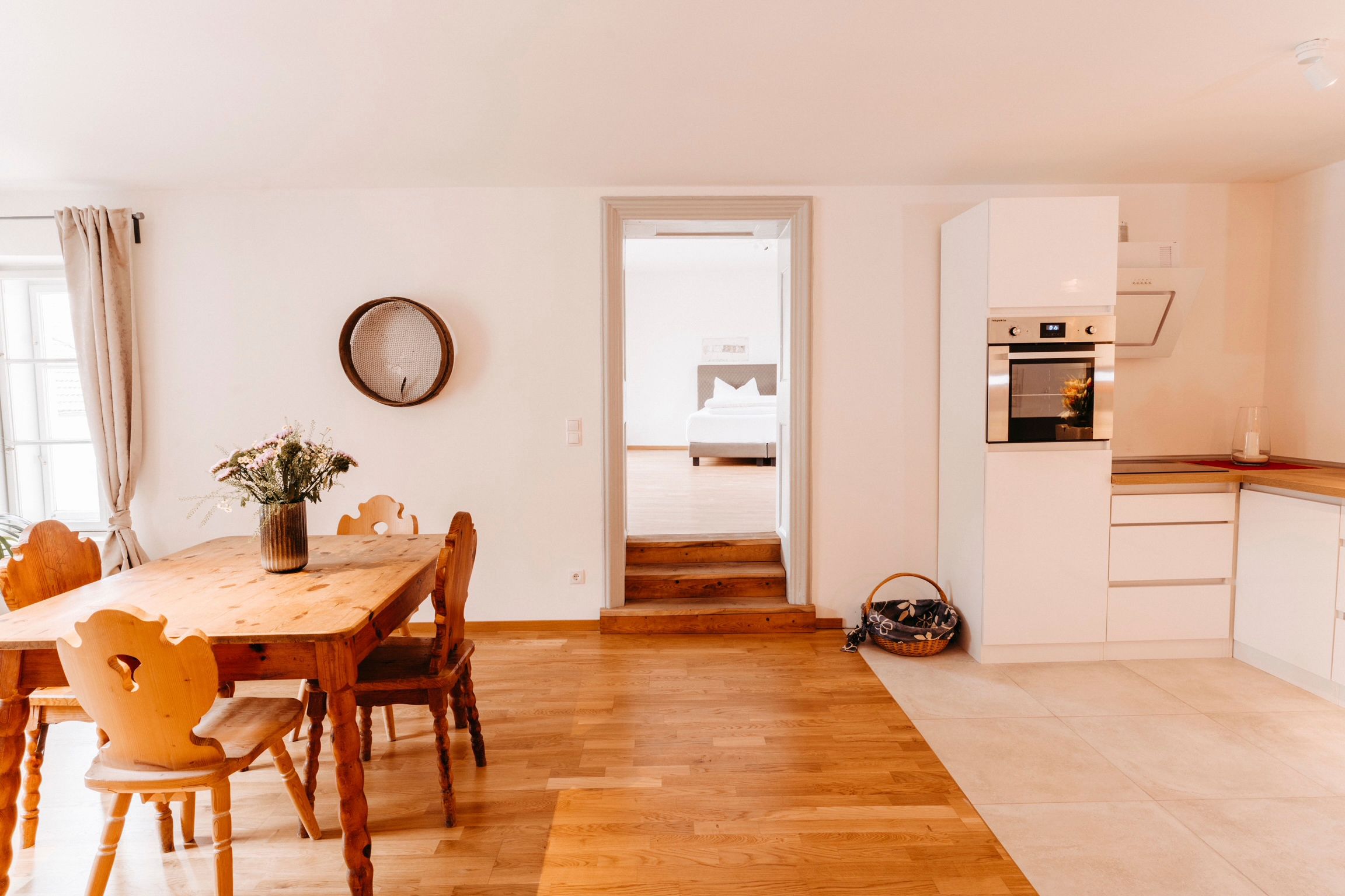 Helle Küche mit Holztisch, Stühlen und modernen weißen Schränken. Durchgang zu einem Schlafzimmer im Hintergrund.