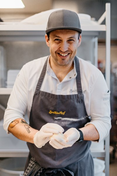 Ein lächelnder Koch mit Schürze und Handschuhen formt Teig in einer Küche.