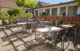 Gastgarten mit Tischen und St&uuml;hlen im Freien, umgeben von Pflanzen und einem Holzzaun.